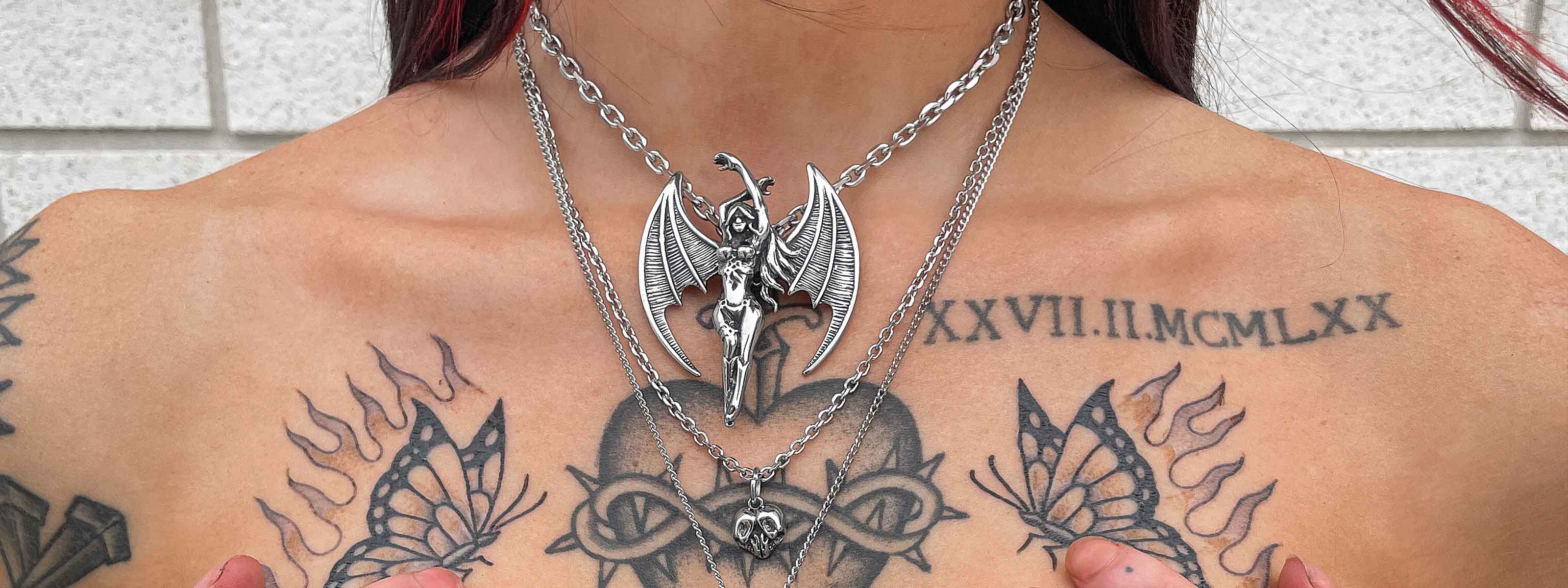 A person with tattoos on their chest wears two silver chain necklaces—one with a winged figure pendant and the other with a smaller, horned skull pendant. Roman numerals and butterfly tattoos are visible.