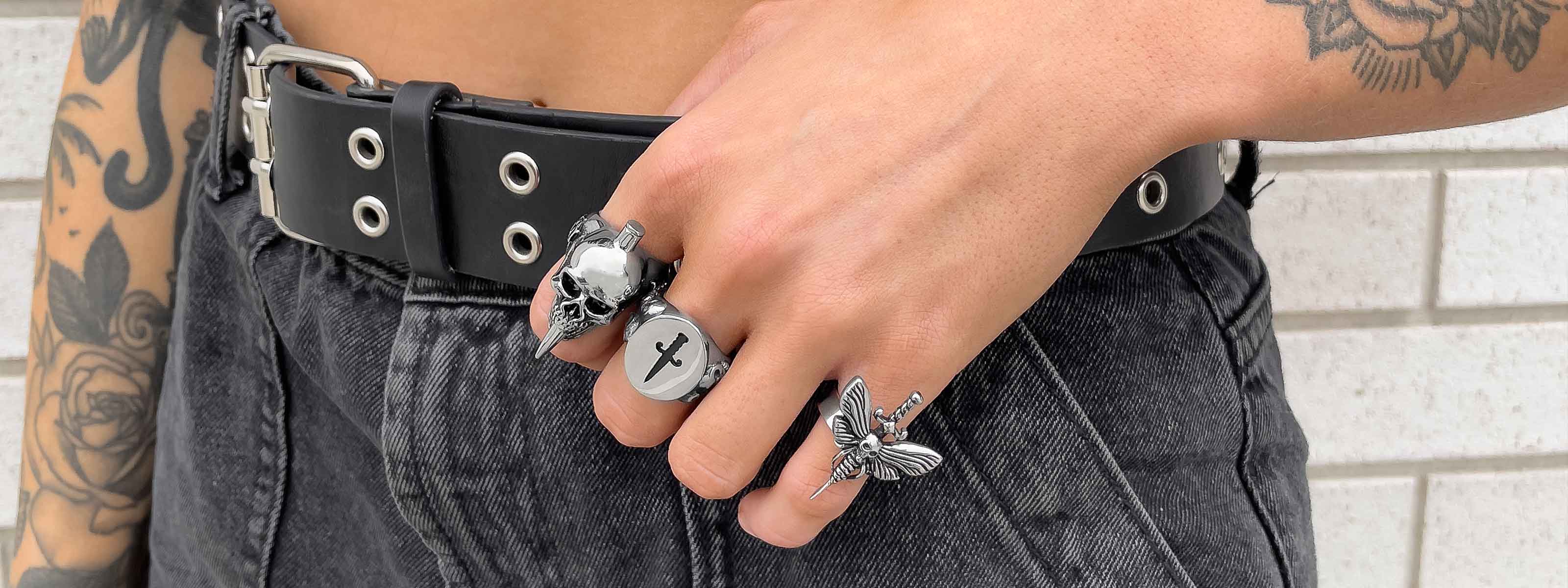 A person with tattoos wears a black belt, high-waisted denim jeans, and silver rings shaped like a skull, a cross, and a moth, standing in front of a tiled white wall.
