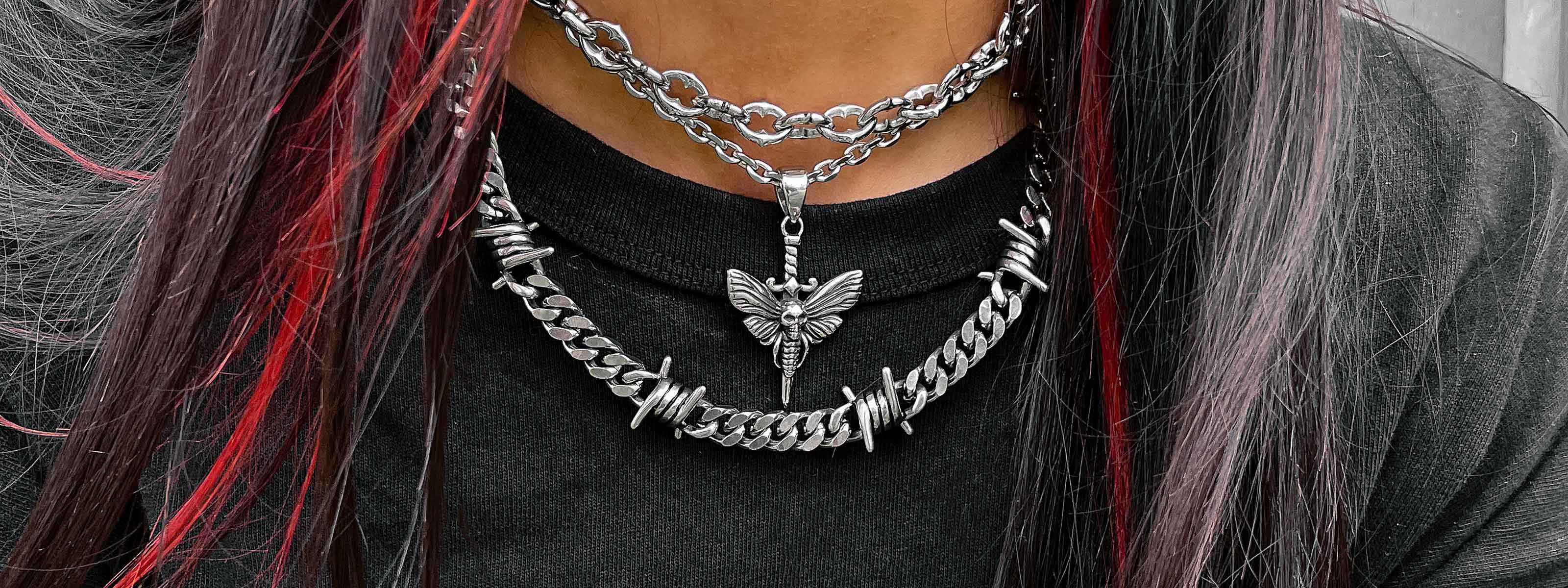 Close-up of a person wearing layered silver chains, one with a moth pendant and another with barbed wire details. The person has long dark hair with red streaks and is wearing a black top.