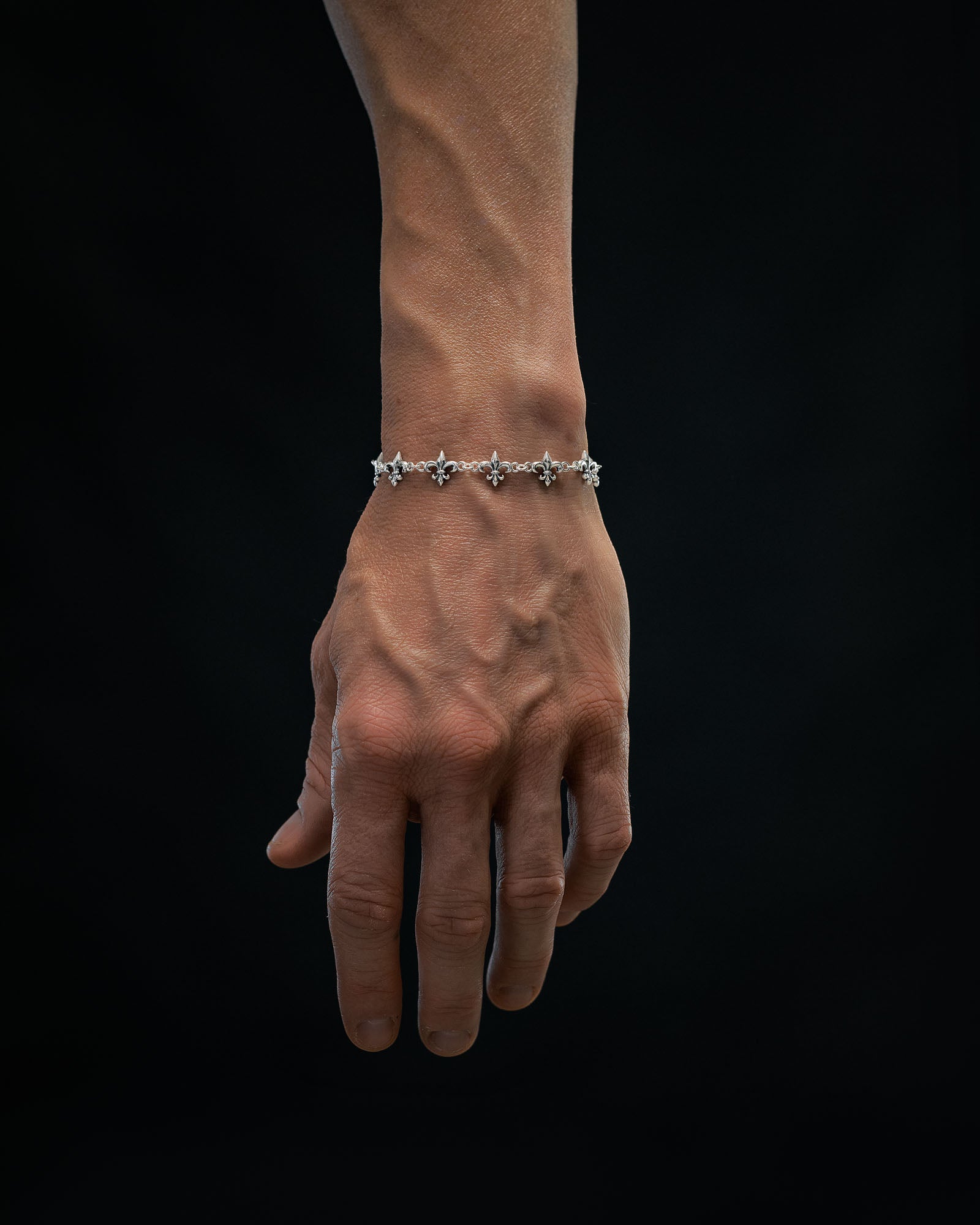 A close-up of a hand with visible veins wearing the Statement Collective 925 Sterling Silver "Fleur De Lis" 10mm Bracelet, set against a solid black background.