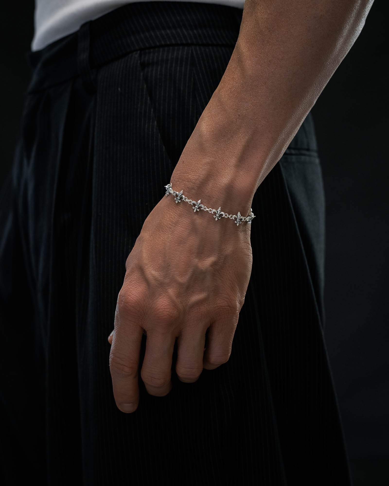 A close-up of a person's hand and wrist adorned with the Statement Collective 925 Sterling Silver "Fleur De Lis" 10mm Bracelet, paired with black pinstriped pants and a white shirt against a dark background.