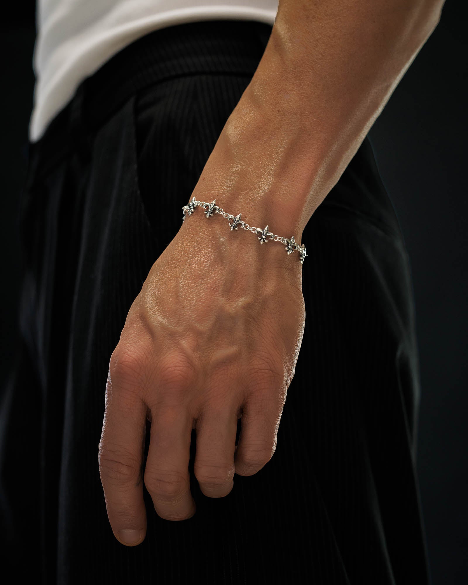 A close-up of a person's hand wearing the Statement Collective 925 Sterling Silver "Fleur De Lis" 10mm Bracelet, featuring intricate designs, set against a background of black pants and a white shirt.
