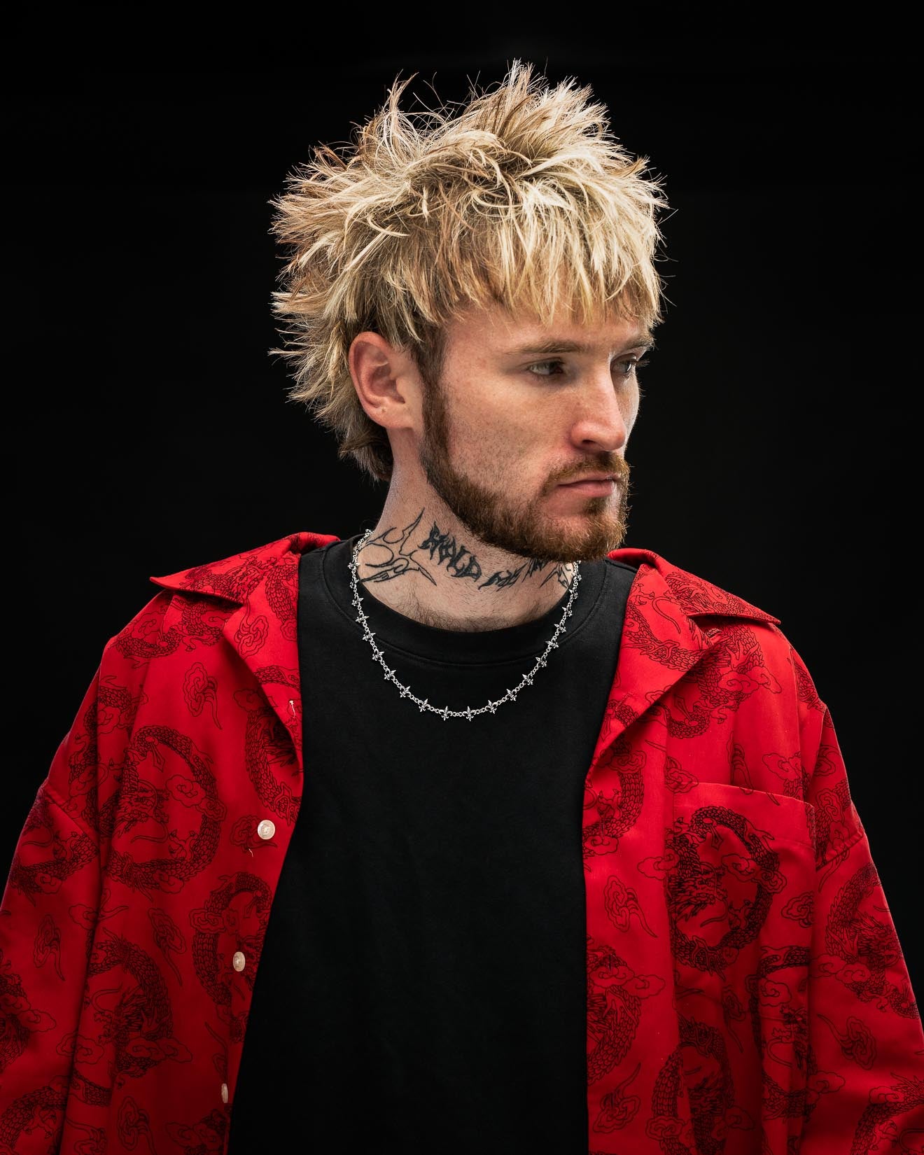 A person with spiky blond hair and a beard wears a red patterned jacket over a black shirt, accessorized with Statement Collective's 925 Sterling Silver "Fleur De Lis" 10mm Chain, and has tattoos on their neck, standing against a black background.