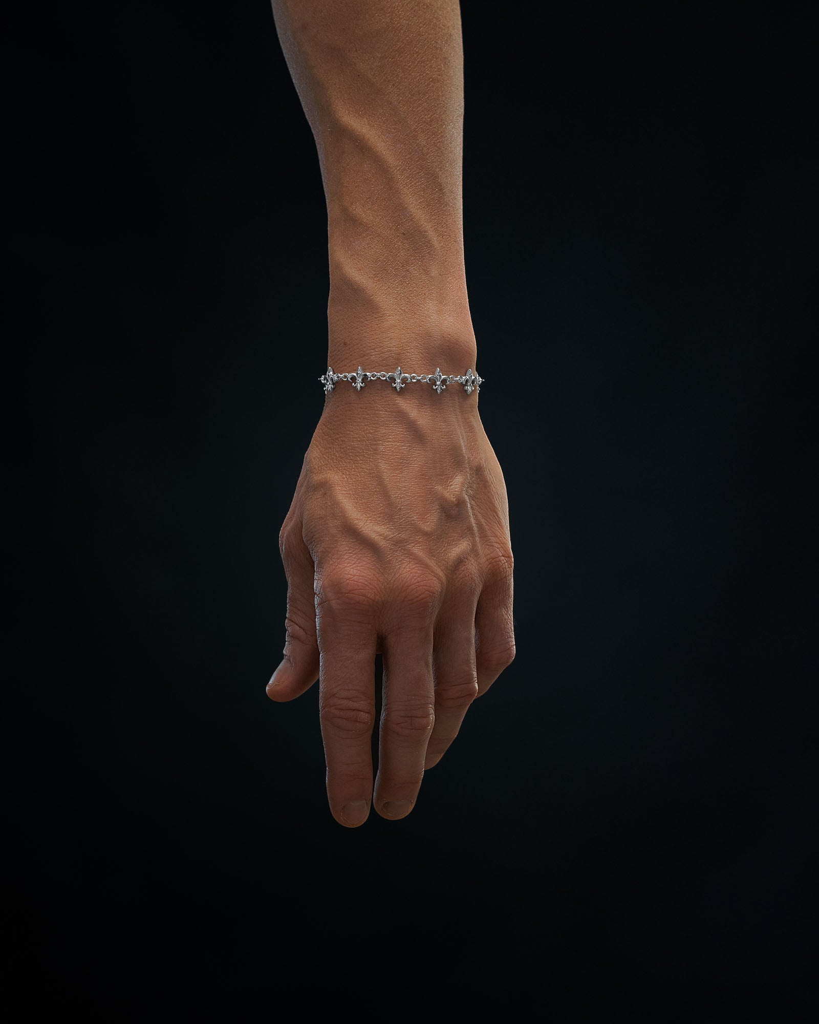 A hand with prominent veins wears the Statement Collective "Fleur De Lis" 10mm Bracelet, a delicate silver piece featuring small floral charms, set against a plain dark background.