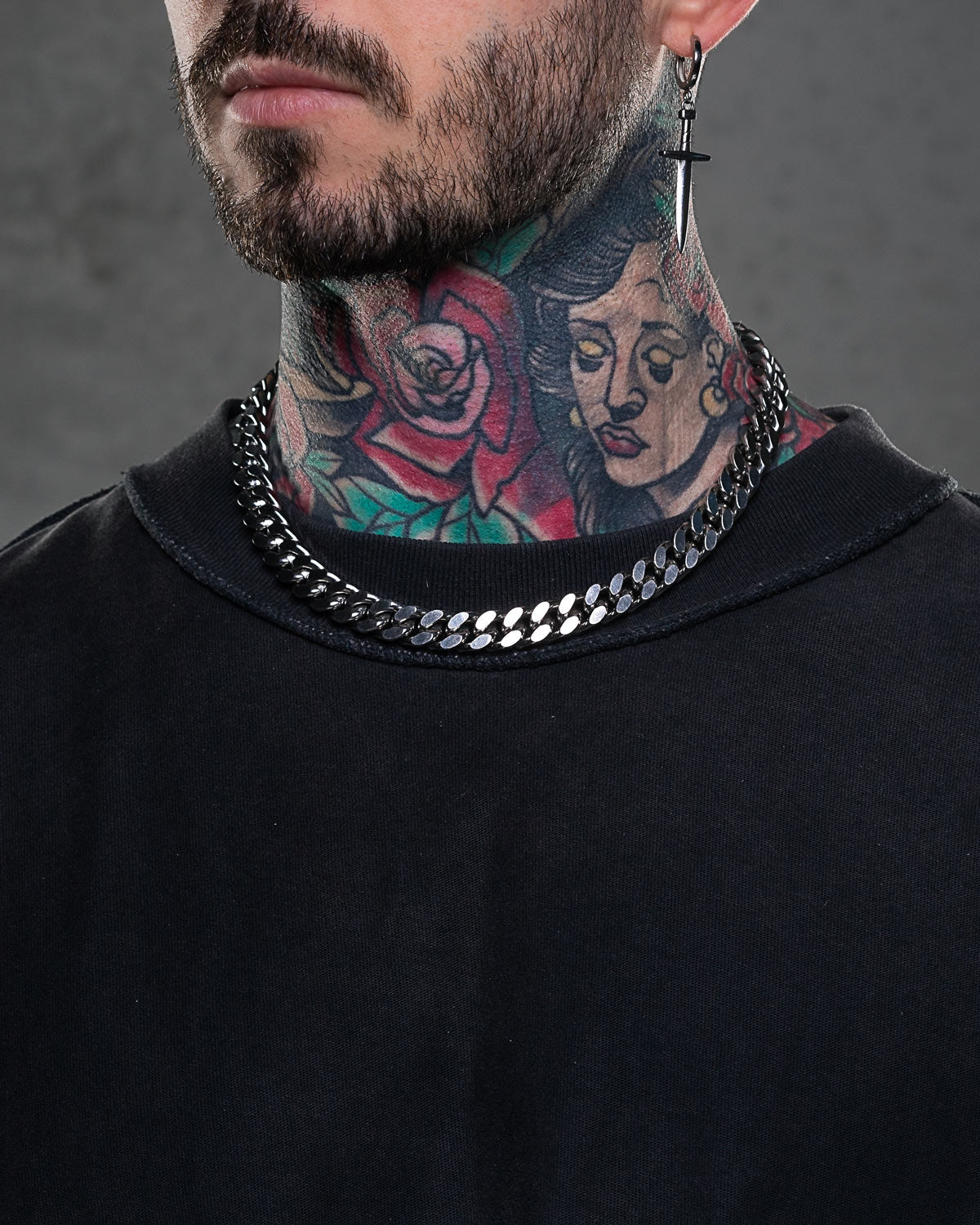A man with a beard and colorful neck tattoos sports a black shirt, 11mm Cuban Chain necklace, and cross earring. A rose and woman's face tattoo accentuate his bold punk style.