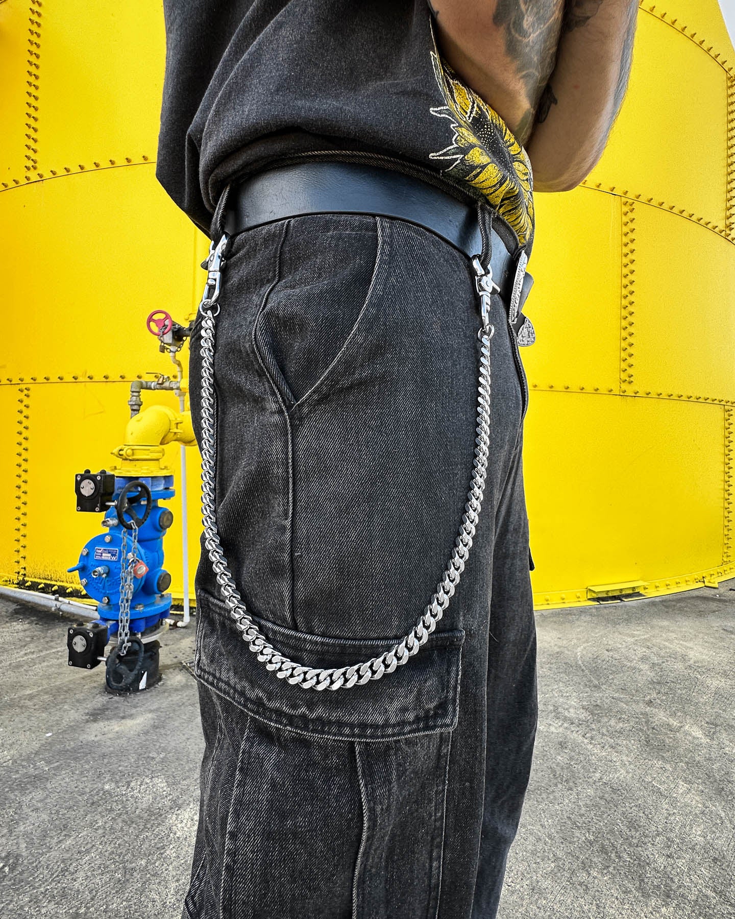 Wearing black jeans and a black shirt with a yellow design, a person stands by a large yellow industrial tank, sporting an 11mm Cuban Wallet Chain. In the background, a blue and yellow valve assembly is visible.