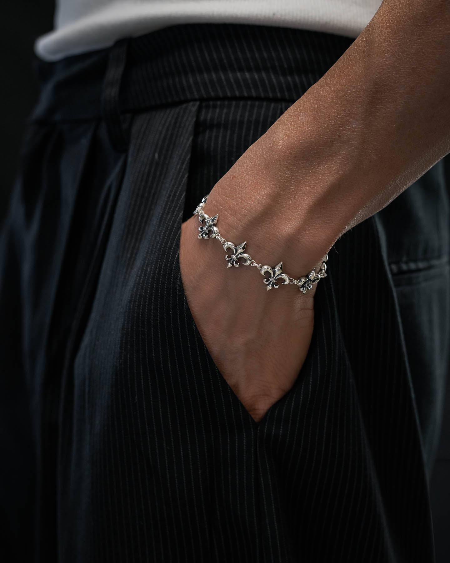 A close-up of a person wearing the Statement Collective 925 Sterling Silver "Fleur De Lis" 15mm Bracelet, with their hand in the pocket of black pinstriped pants and dressed in a white shirt.