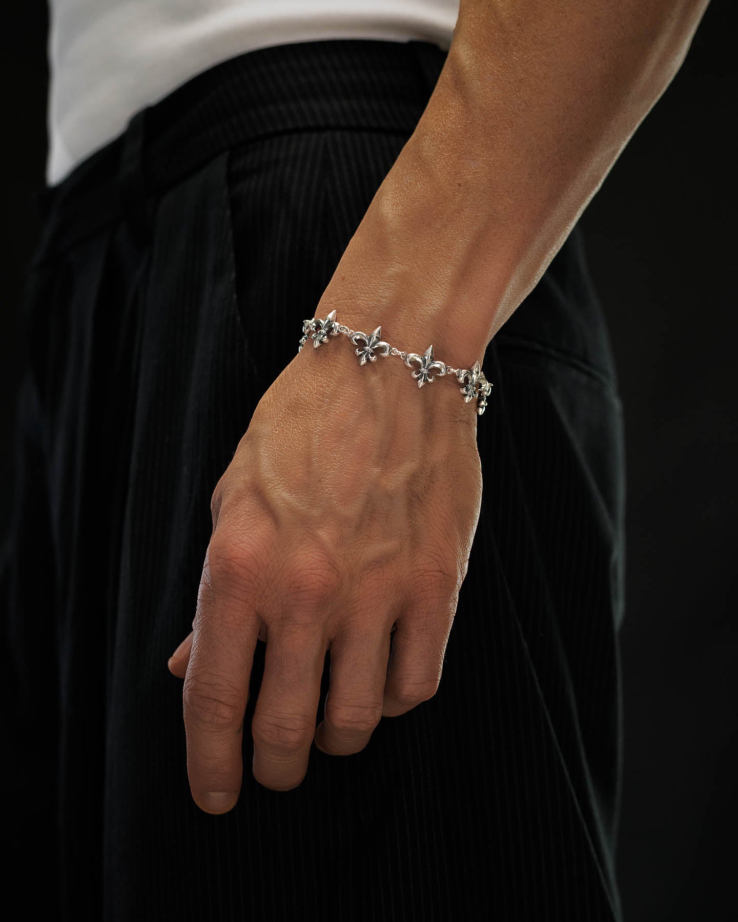 A close-up of a person's hand and wrist adorned with the Statement Collective 925 Sterling Silver "Fleur De Lis" 15mm Bracelet. They are dressed in a white shirt and black pinstripe pants, set against a dark background.