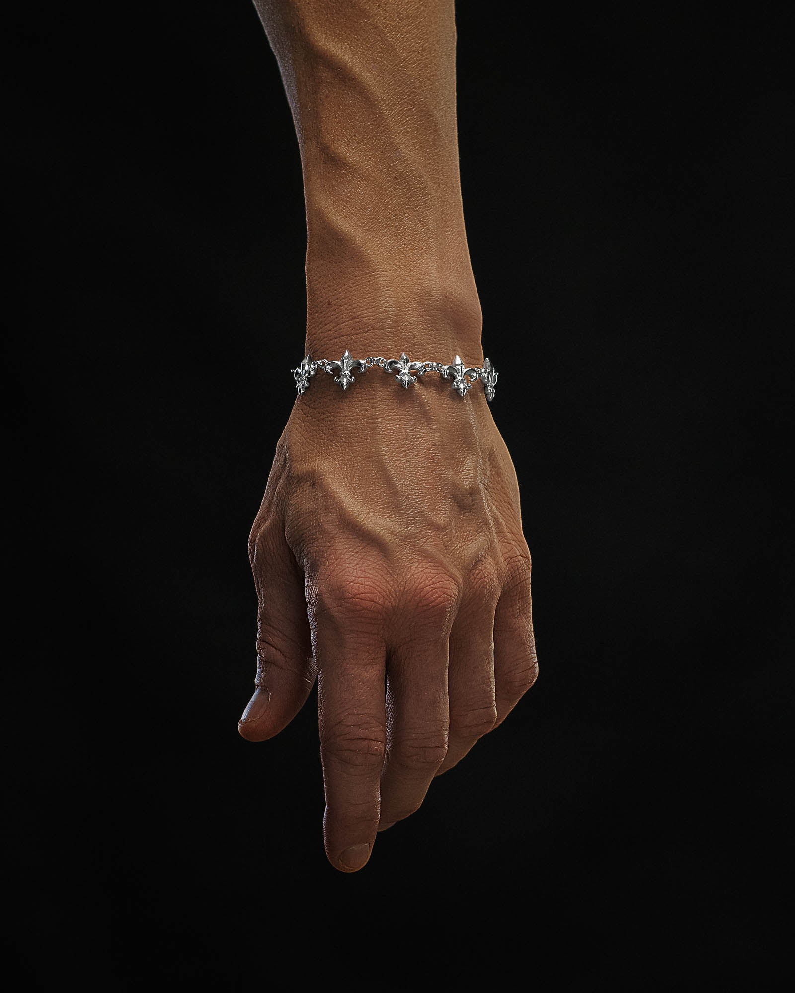 A hand with pronounced veins wears the Statement Collective "Fleur De Lis" 15mm Bracelet, its repeating fleur-de-lis design in silver set against a solid black background.