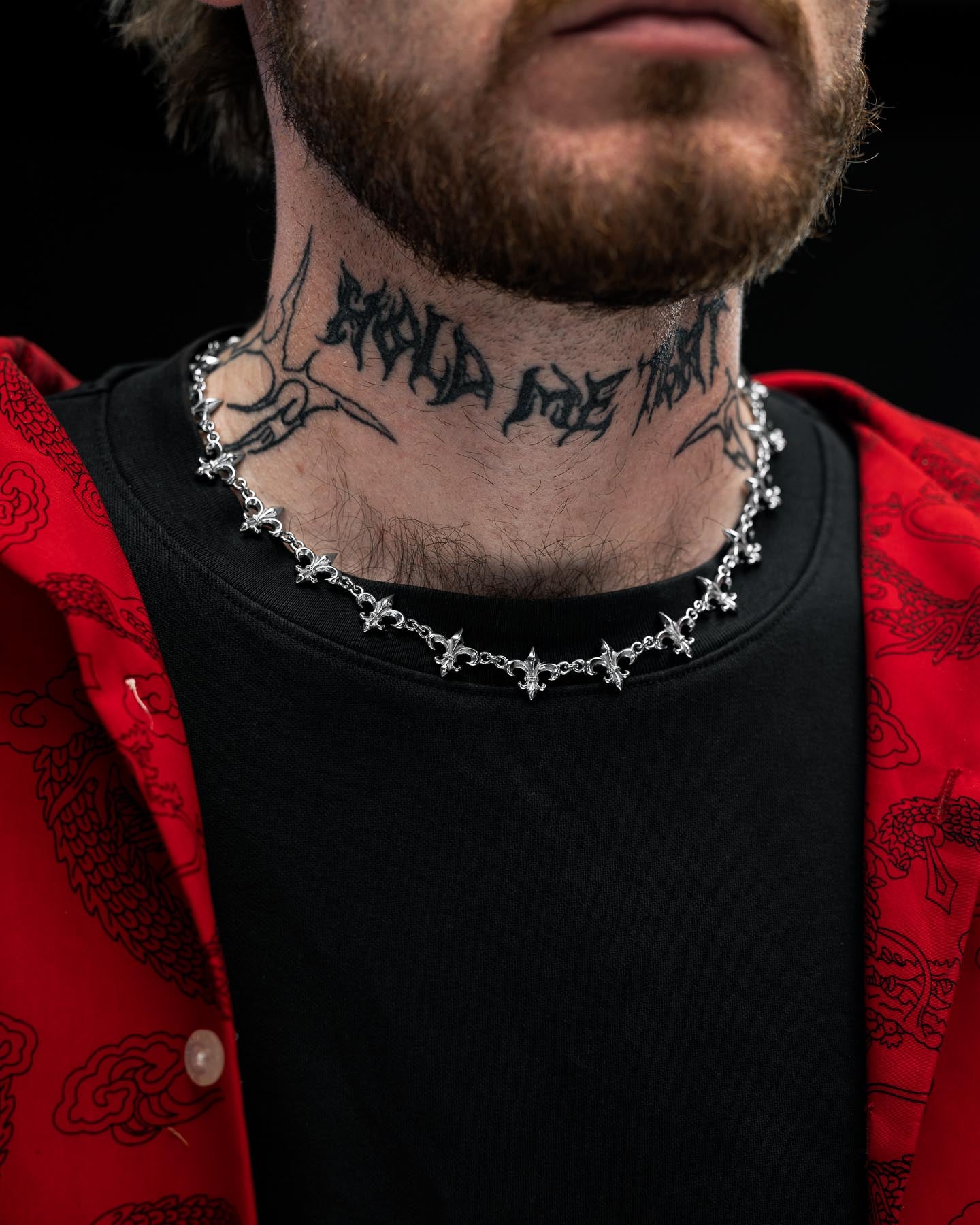 A person with a beard and mustache wears a black shirt, red jacket, the "Fleur De Lis" 15mm Chain by Statement Collective, and has a tattoo with decorative script on their neck. The background is dark.