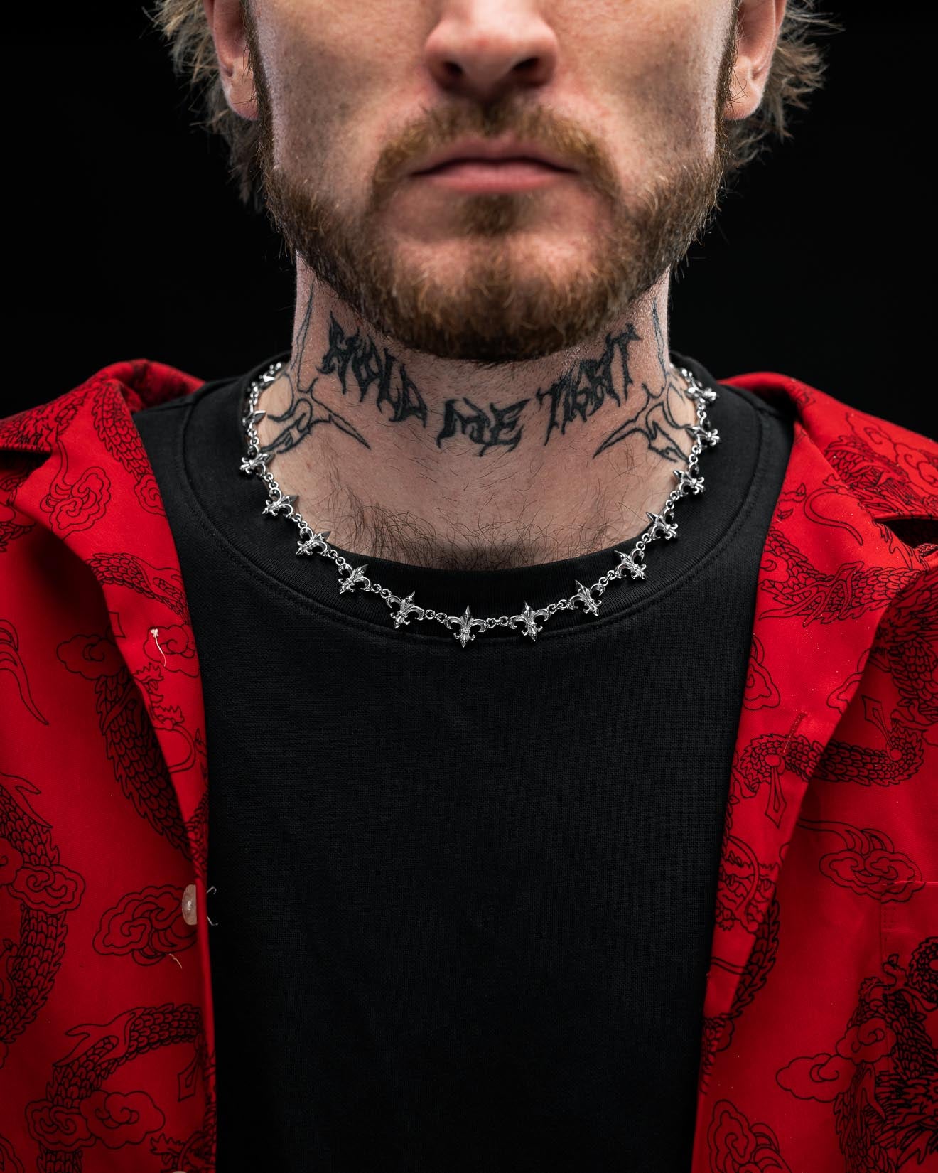 A man with a beard wears the Statement Collective "Fleur De Lis" 15mm Chain, a black shirt, and a red patterned jacket. He has a large tattoo with unreadable text across his neck, set against a dark background.