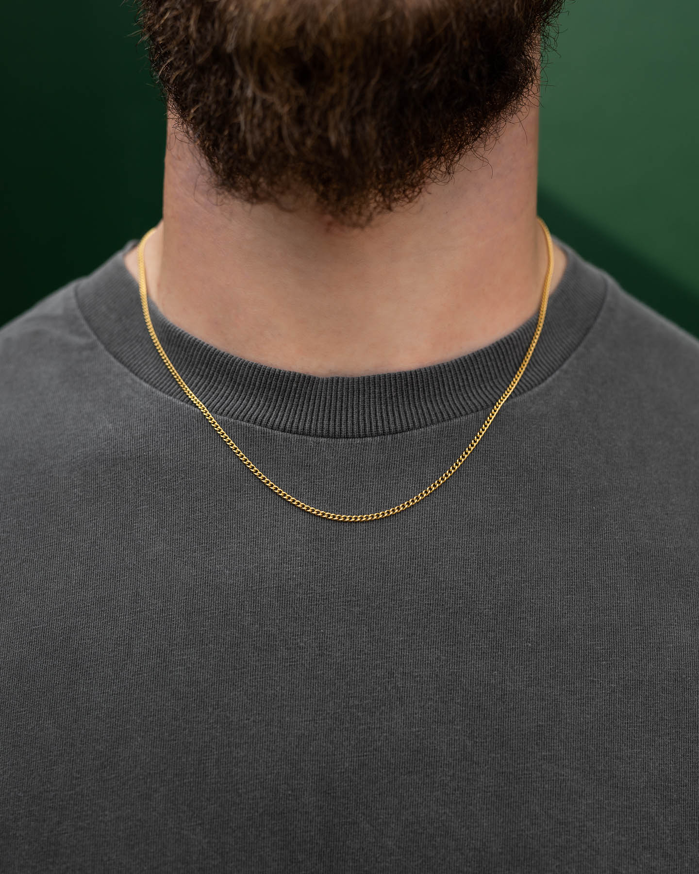 A close-up of a person in a gray crew-neck shirt wearing the 2.3mm Curb Chain (Gold) necklace, featuring a thin gold plated stainless steel design, with a short brown beard and a green background.
