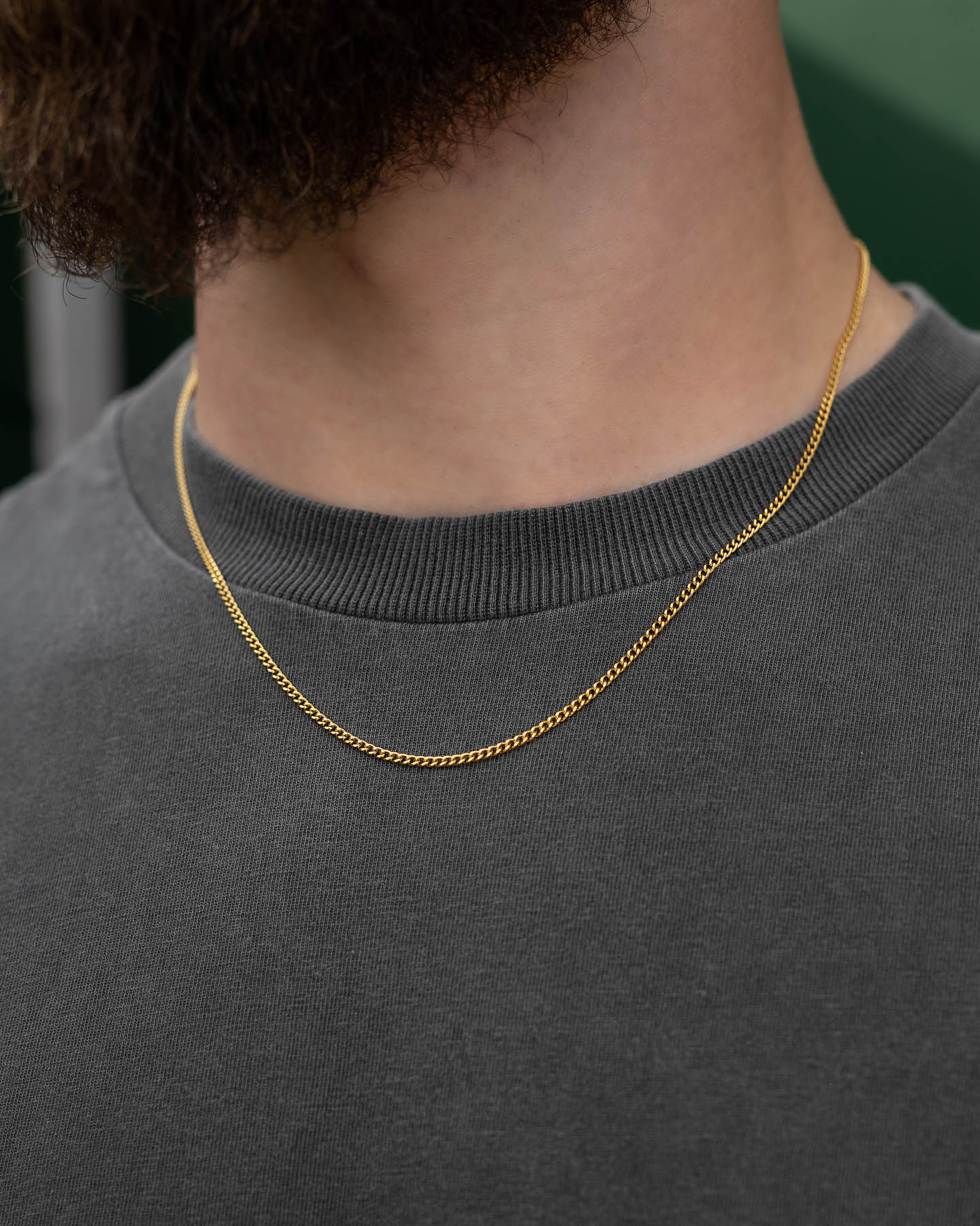 Close-up of a person wearing the 2.3mm Curb Chain (Gold) over a dark grey crew-neck shirt, with part of a beard visible at the top of the image.