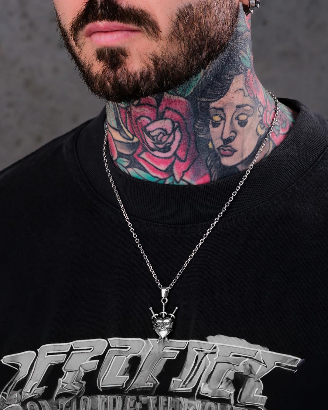 A person with colorful rose and portrait neck tattoos wears the "3 of Swords" Pendant—a heartbreak necklace with a crown charm—and a black shirt with metallic text. Only the lower half of their face is visible.