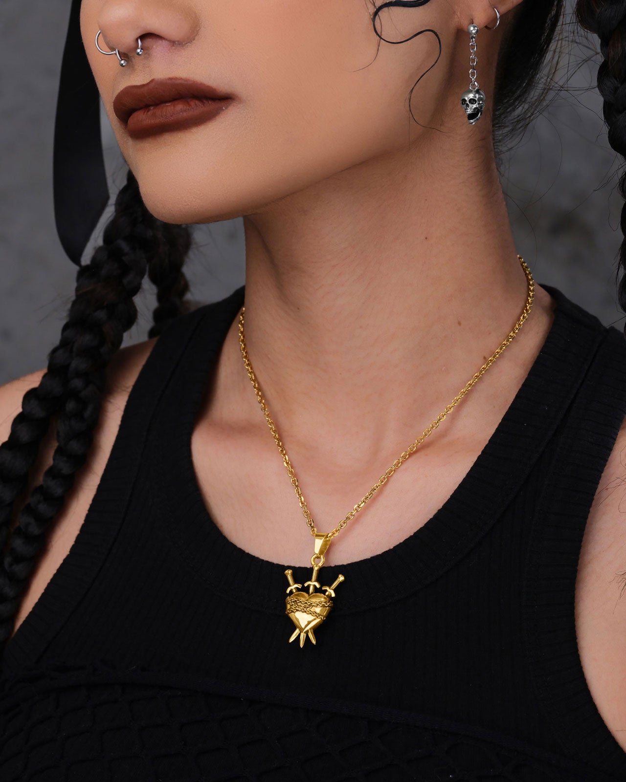 A person with braided hair and facial piercings wears a black sleeveless top, the striking "3 of Swords” Pendant (Gold), a silver skull earring, and has dark brown lips.