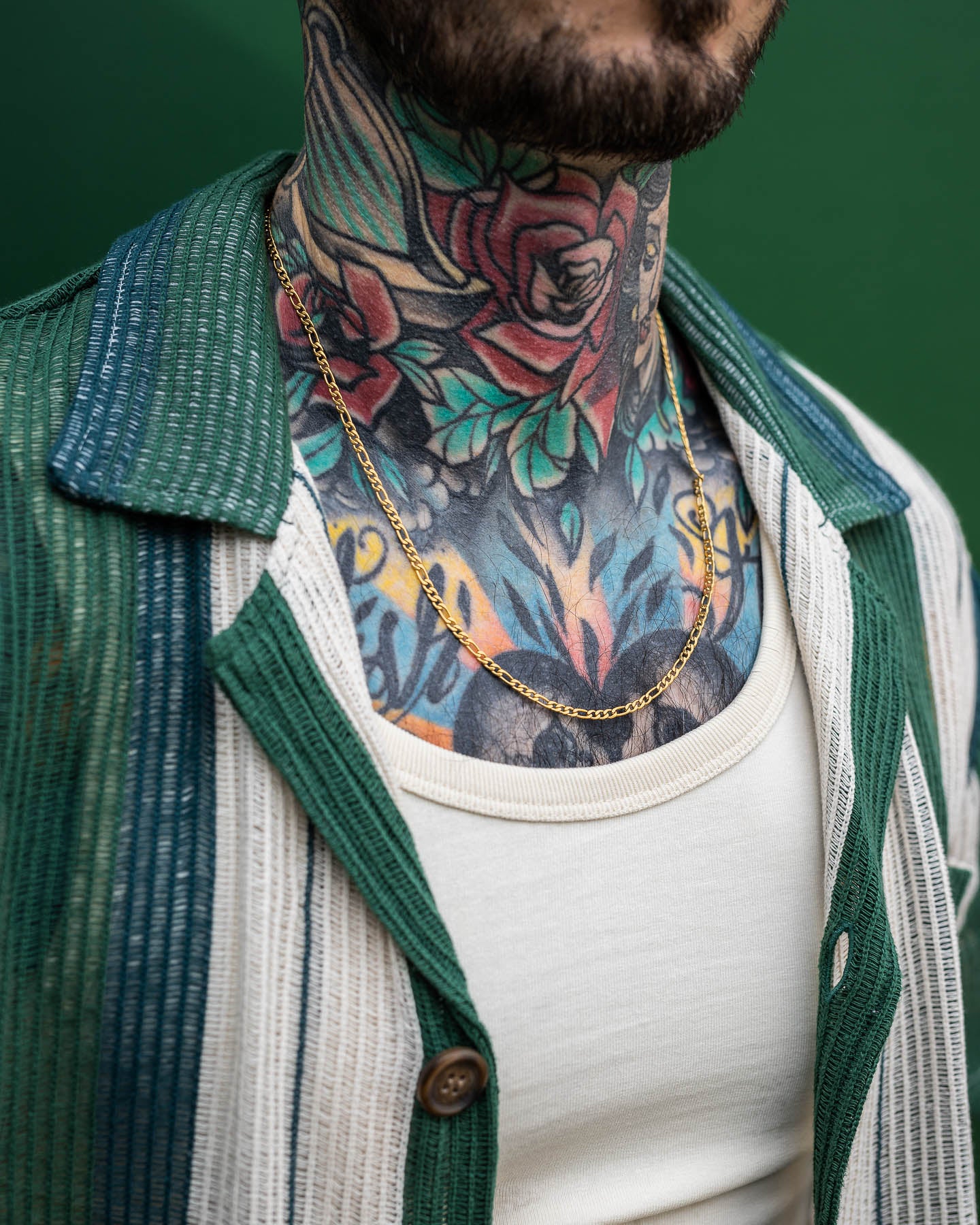 A person with colorful neck and chest tattoos, wearing a green and white shirt over a white tank top, is showcasing the 2.9mm Figaro Chain (Gold) necklace. The background is green.