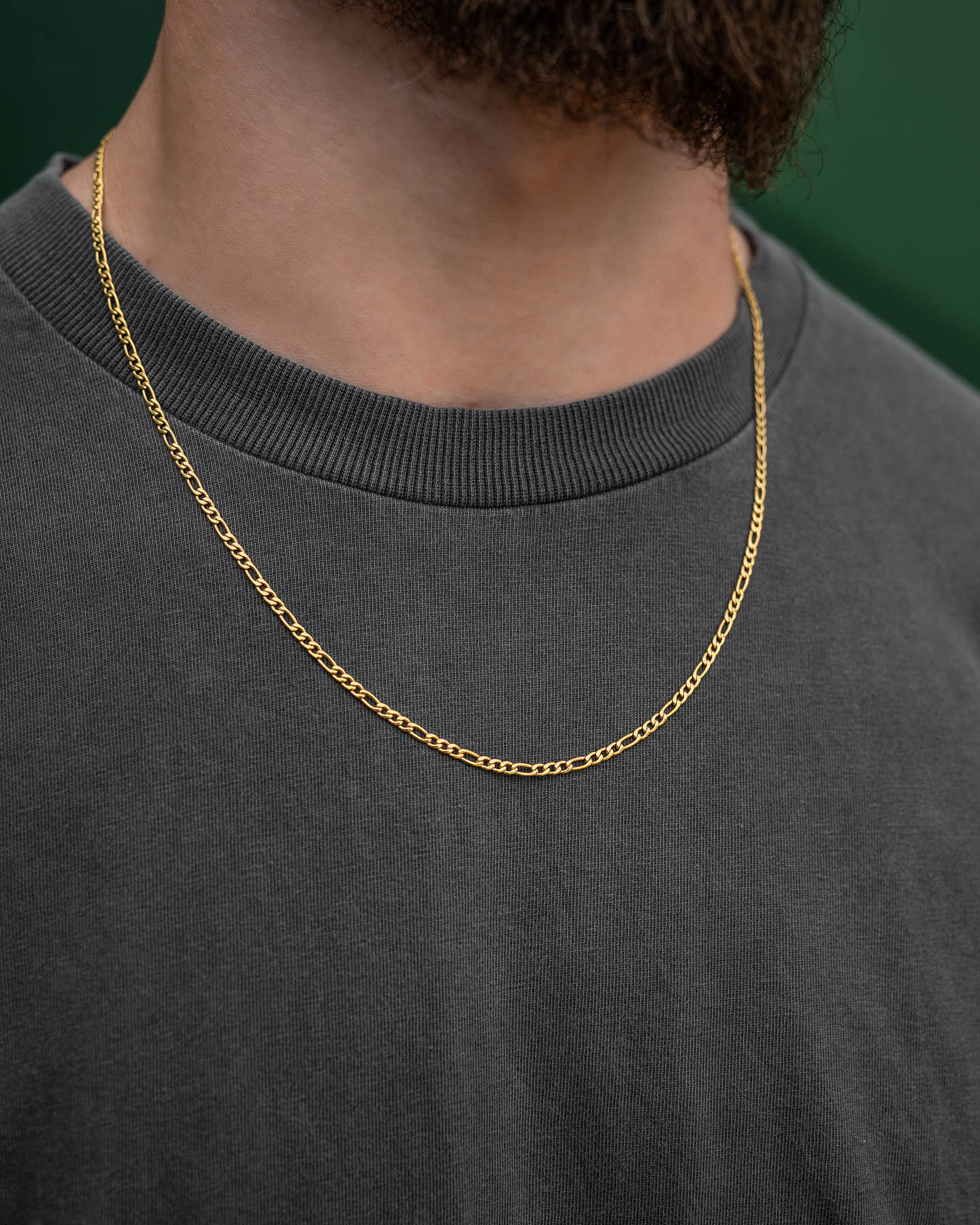 A close-up shows a person wearing a plain dark gray t-shirt and the 2.9mm Figaro Chain (Gold), with part of their beard visible against a green background.