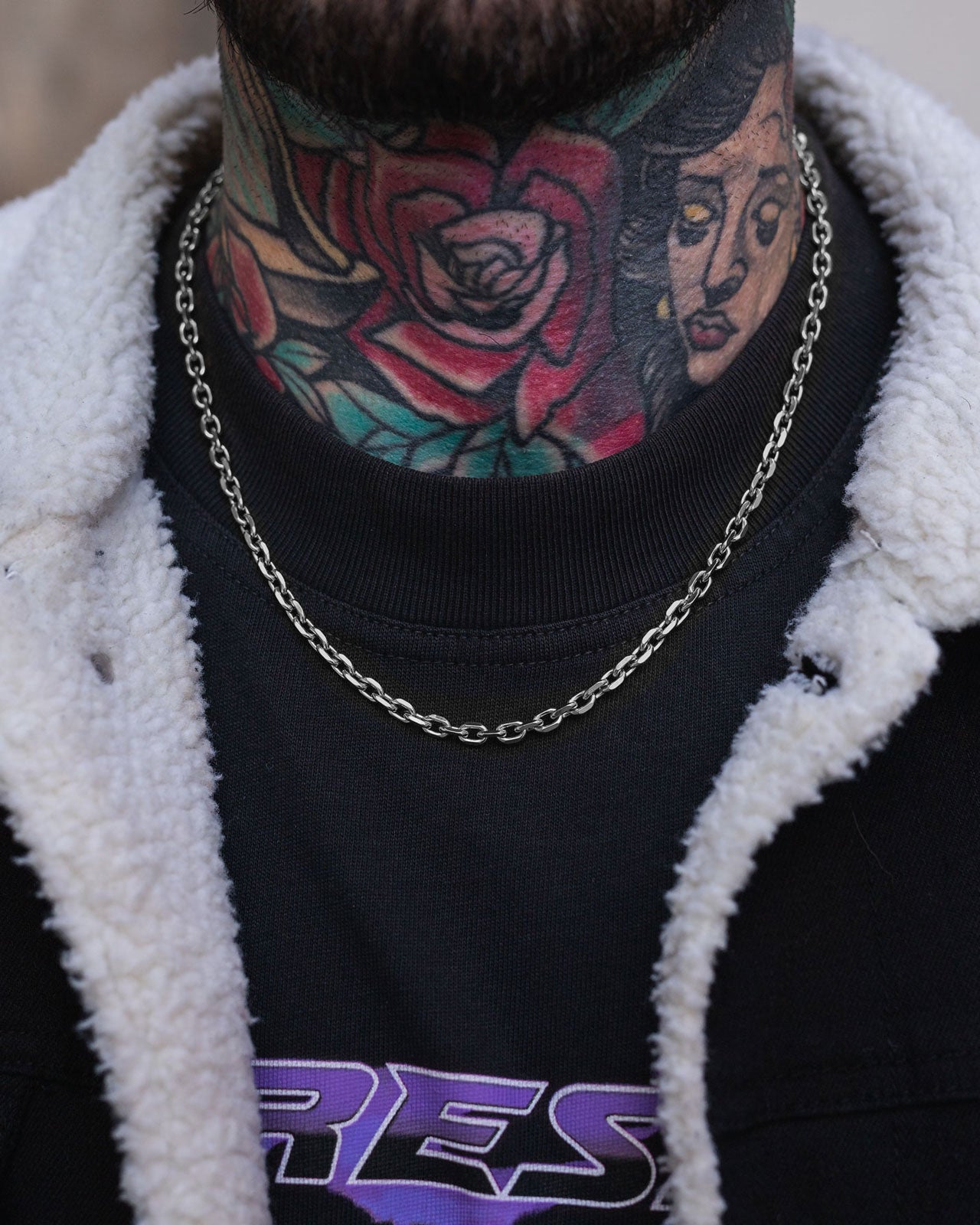 A close-up of a person's tattooed neck, featuring a rose and woman's face, wearing Statement Collective's 4.5mm Cable Chain necklace, a black T-shirt with purple lettering, and a black jacket with a white fleece collar.