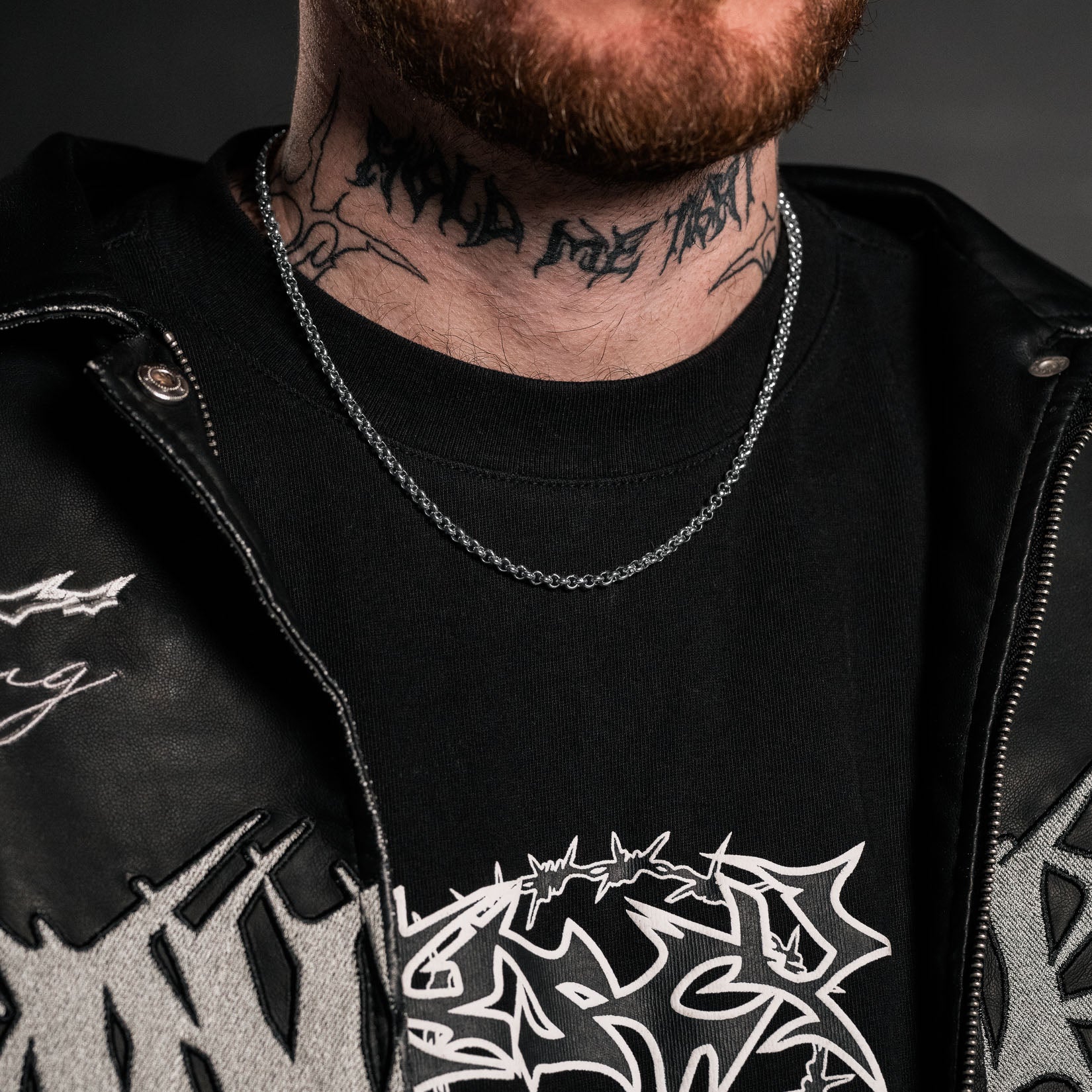 Person wearing a black jacket with white designs and text, featuring a tattoo on the neck. Silver rolo chain necklace by statement collective