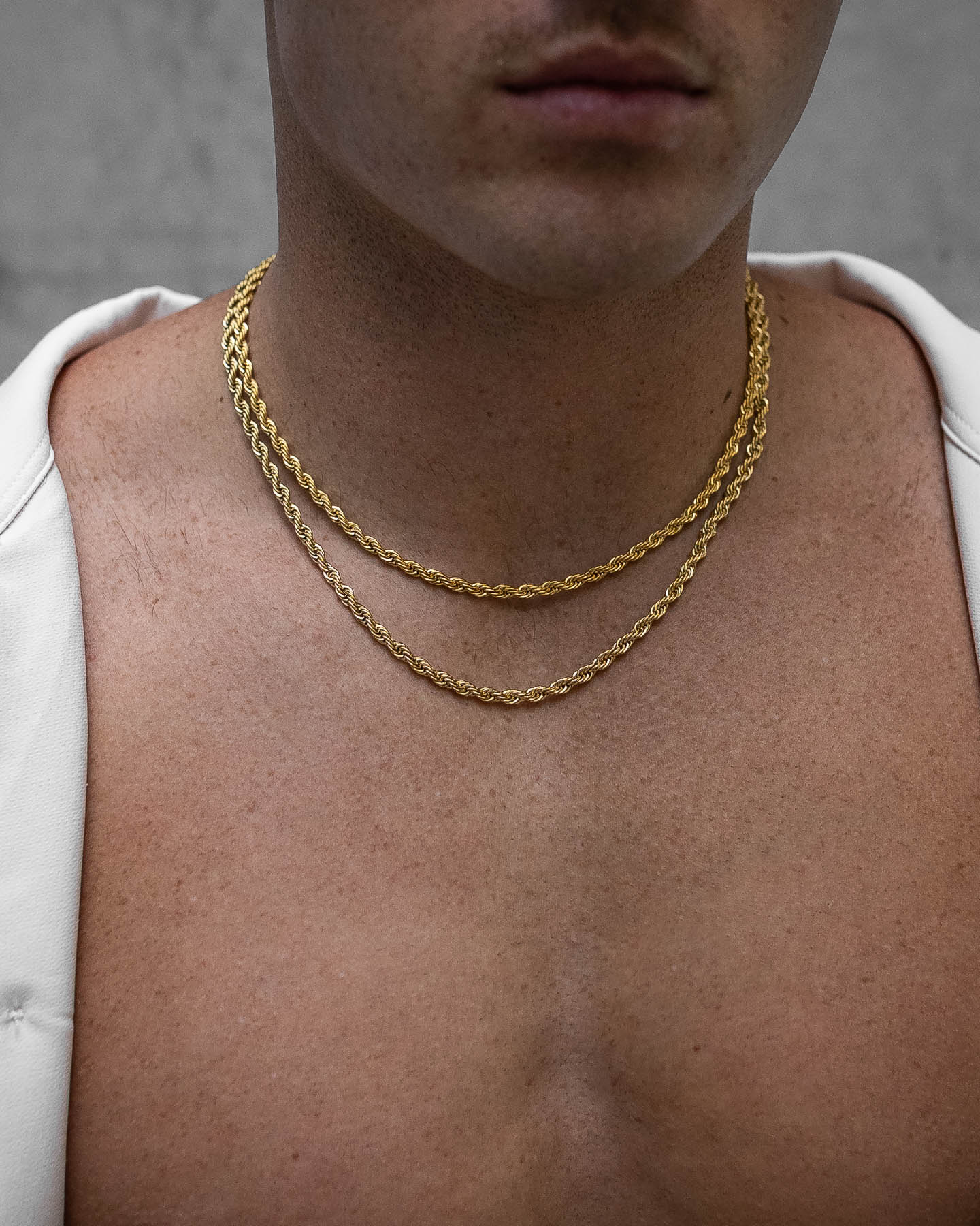 A close-up features a man's chest and neck wearing the 4mm Rope Chain (Gold). A white garment drapes over his shoulders, partially covering his bare chest, while the neutral background is softly blurred.