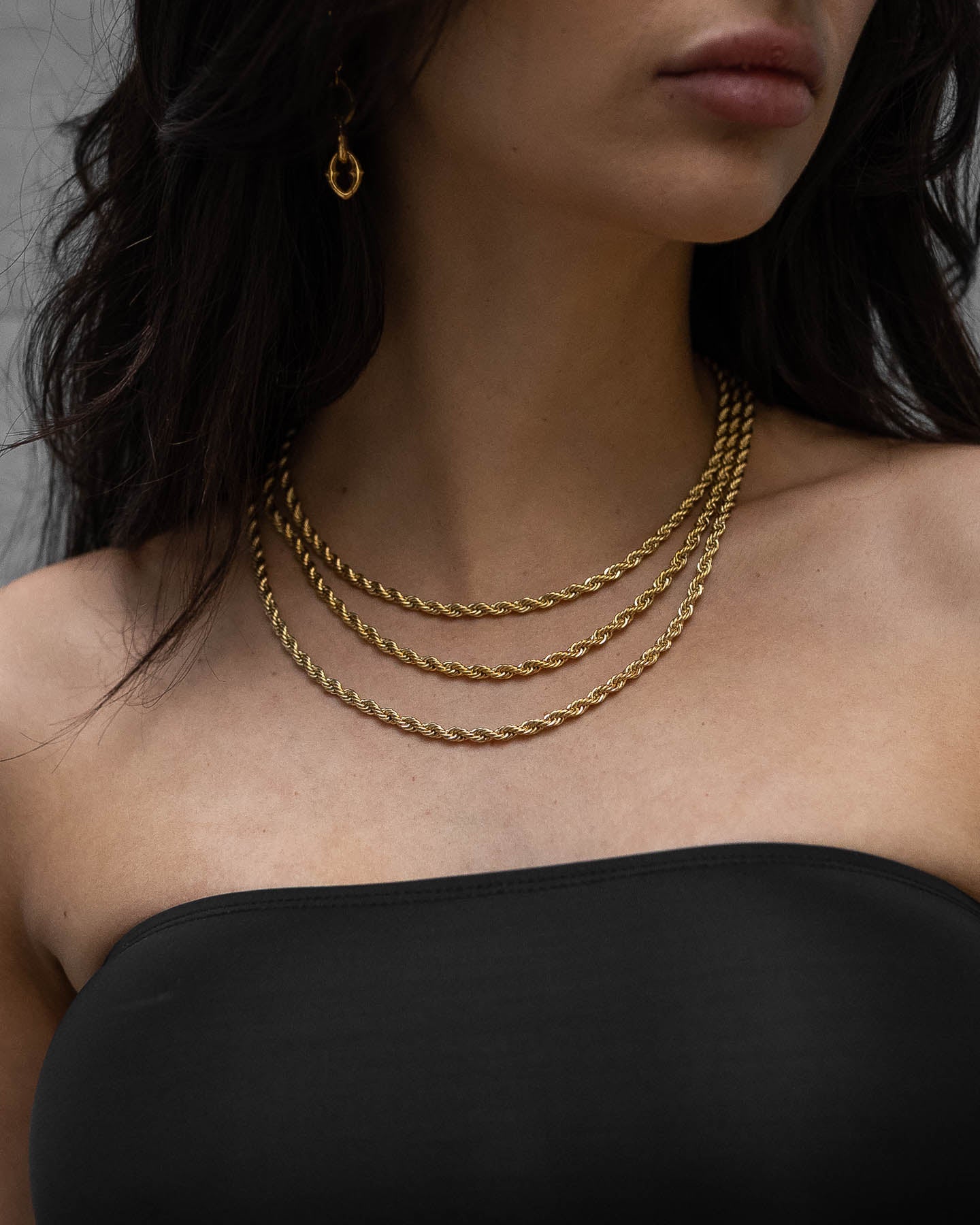 A woman in a strapless black top wears layered gold necklaces and a gold hoop earring. Her dark, wavy hair partly covers her face, drawing attention to the elegance of the 4mm Rope Chain (Gold) around her neck.