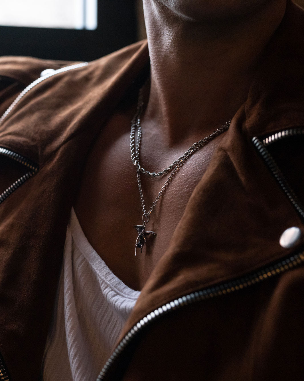 A person styled in a brown suede jacket, white ribbed shirt, and classic jewelry—featuring Statement Collective's 4mm Rope Chain layered with a silver pendant—stands against a dark, softly lit background.