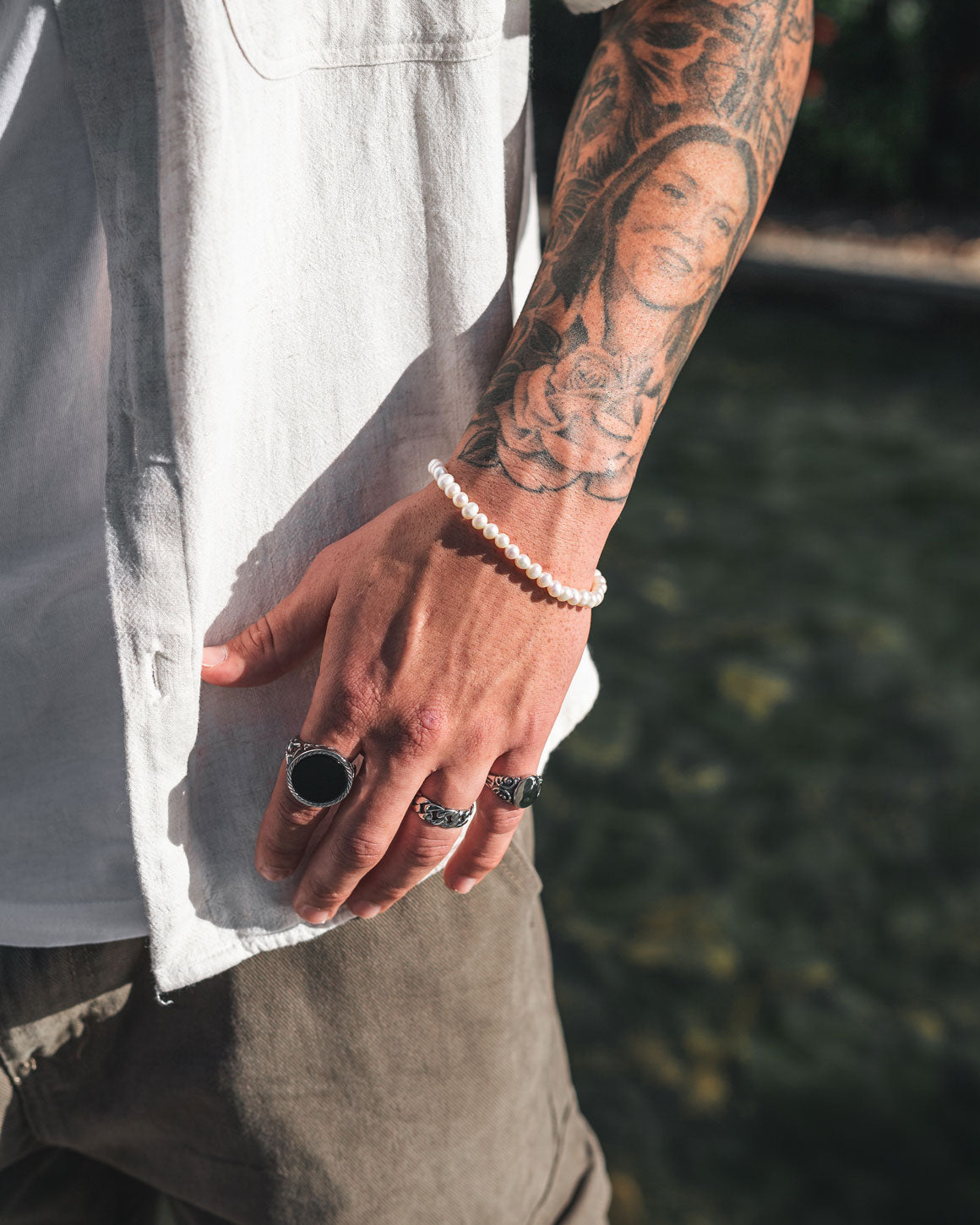 A tattooed arm with a rose and portrait design wears a white shirt, dark pants, silver rings, and the 5mm Freshwater Pearl Bracelet in sunlight over a blurred outdoor background.