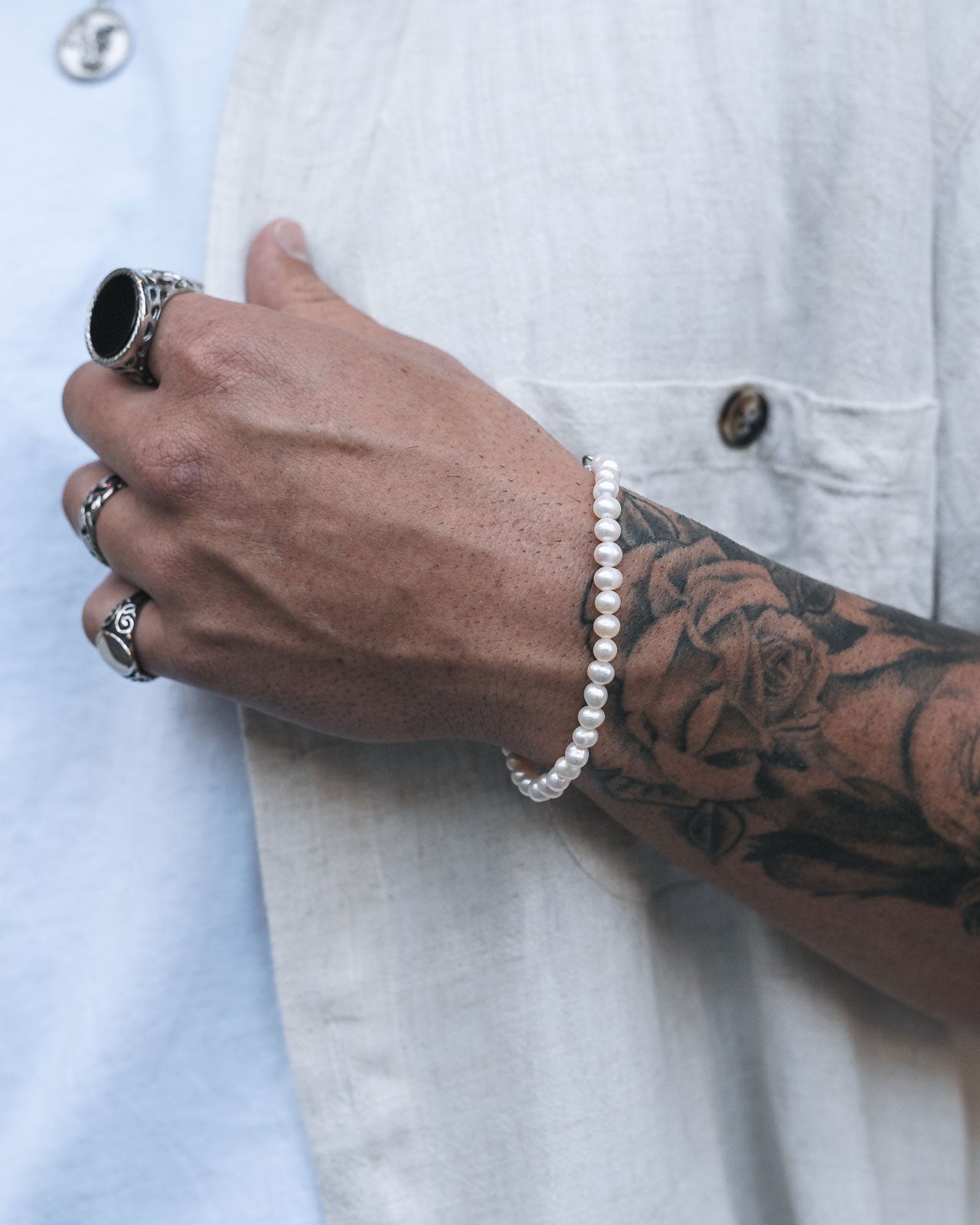 A person with a tattooed forearm and silver rings wears a 5mm Freshwater Pearl Bracelet, paired with a white shirt and light blue t-shirt. Their relaxed hand gently touches the shirt near a button.