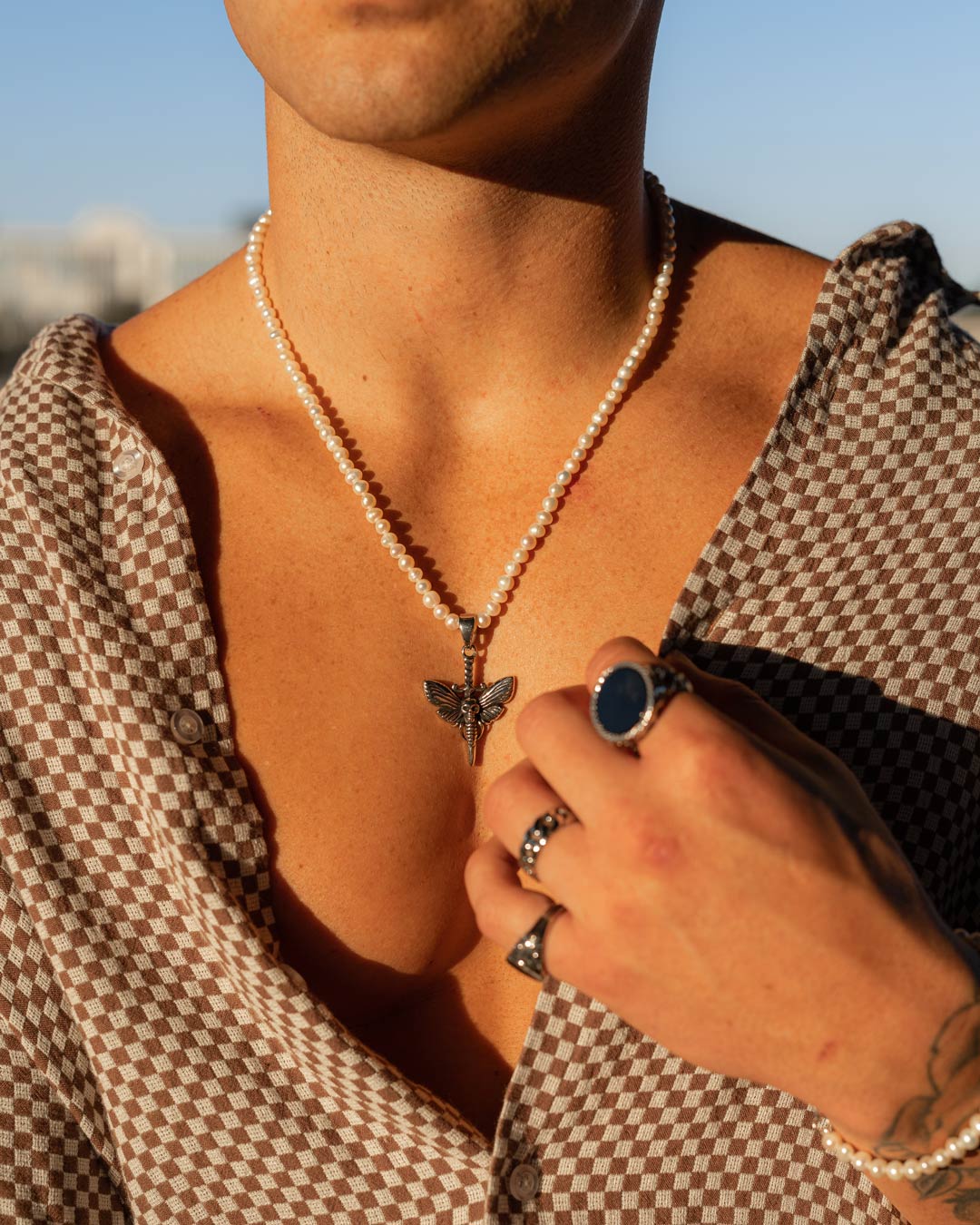 A tanned person wears a chest-open checkered shirt, showing off a Freshwater Pearl Necklace With Custom Pendant Choice and several silver rings, one featuring a dark blue stone.
