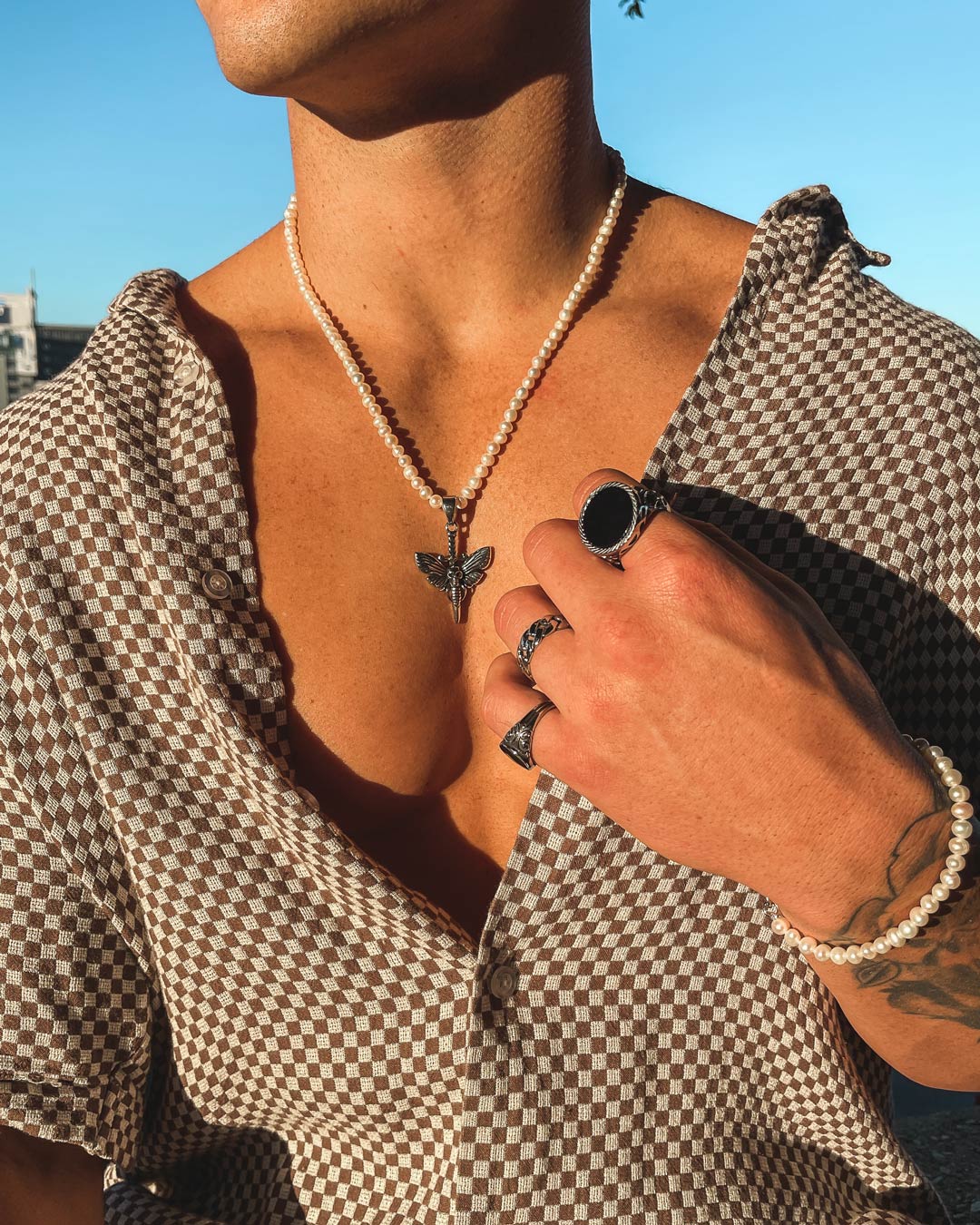 Standing in sunlight, a person in a brown and white checkered shirt—partially unbuttoned to show a Freshwater Pearl Necklace With Custom Pendant Choice—wears several rings, a bracelet, and displays a forearm tattoo.