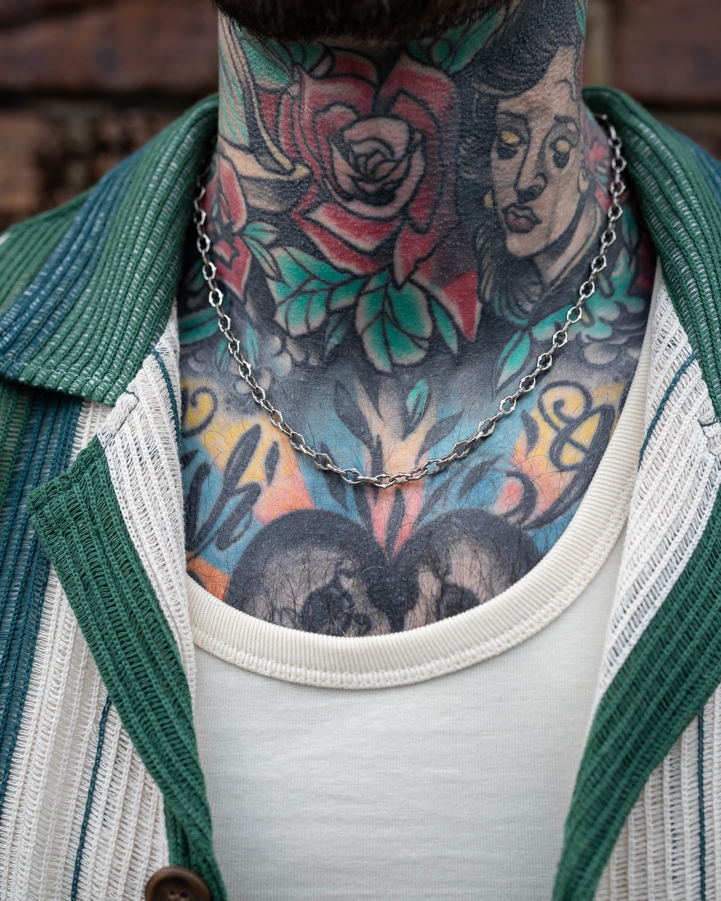 A person in a green and white jacket and white shirt, showing colorful tattoos on their neck and chest, accessorized with "The Cathedral" 6mm Spiked Chain—a bold stainless steel necklace.