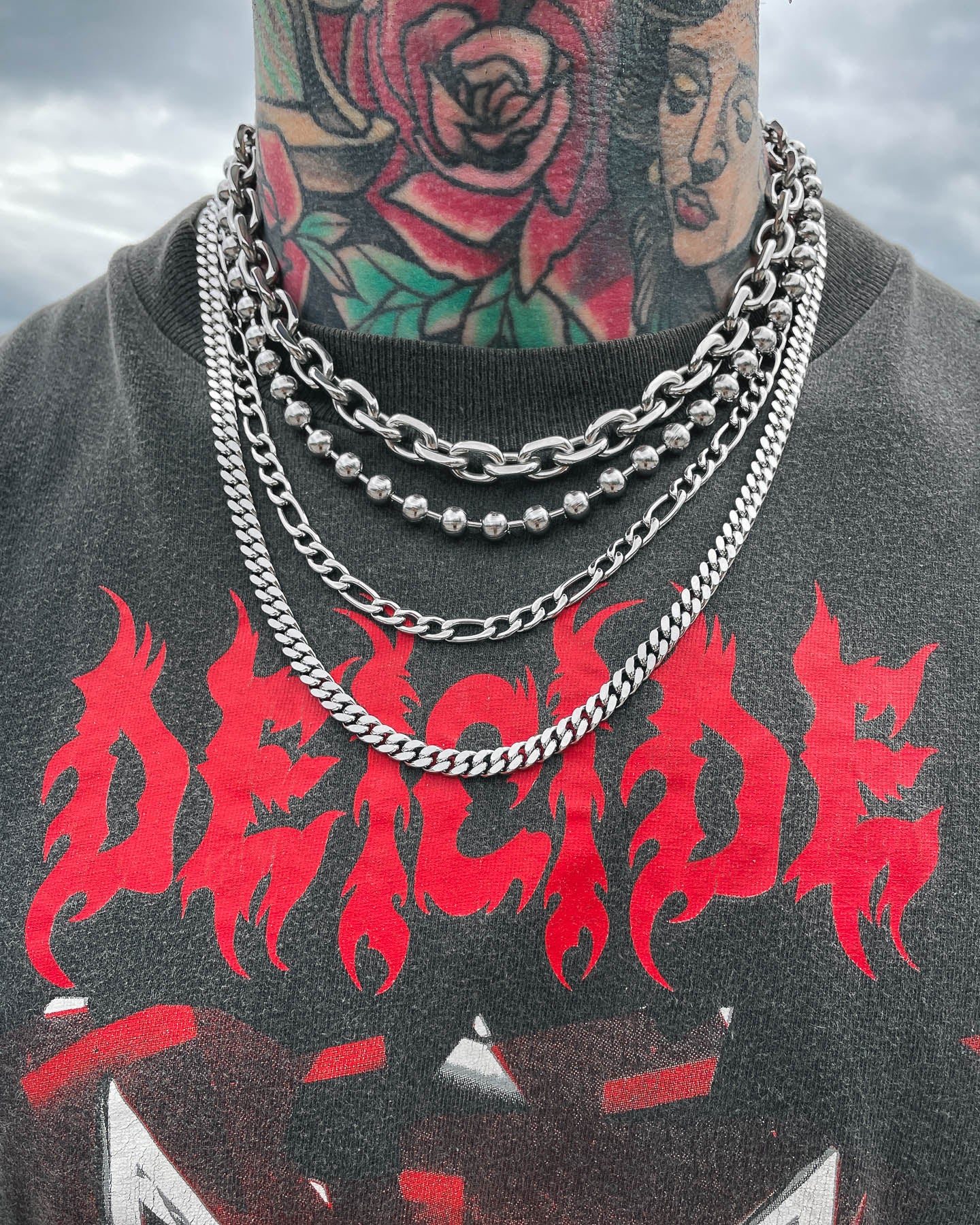 A person wearing layered silver chains, including a 6mm Cuban Chain, and a black shirt with DEICIDE in bold red letters. Their tattooed neck—featuring a rose and a woman's face—stands out against the cloudy sky.