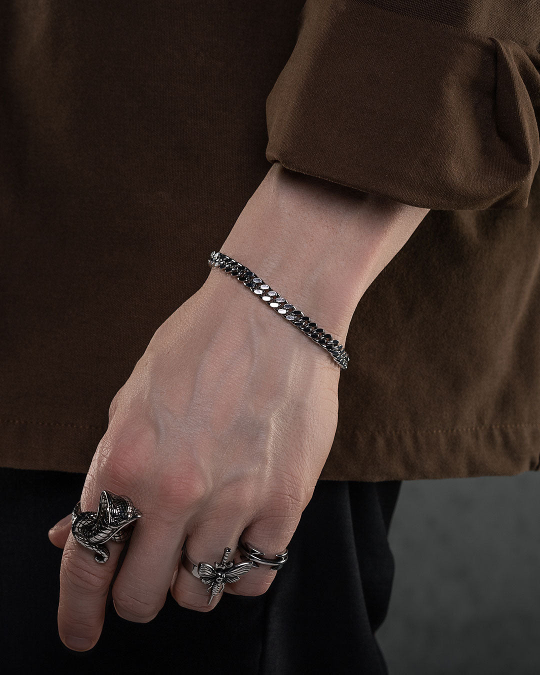 A person in a brown long-sleeve shirt shows their left hand wearing the 6mm Cuban Bracelet and two ornate silver animal-shaped rings, set against a dark, blurred background.