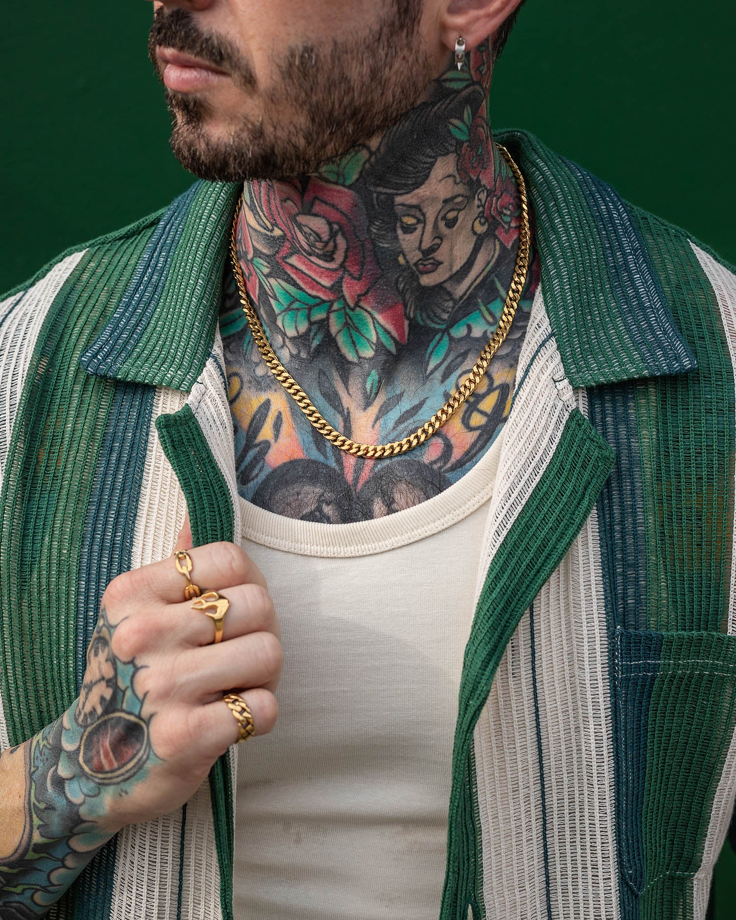 A man with colorful neck tattoos and gold rings wears a green and white striped shirt over a tank top, holding the shirt open to display his 6mm Cuban Chain (Gold) necklace and jewelry against a dark green background.