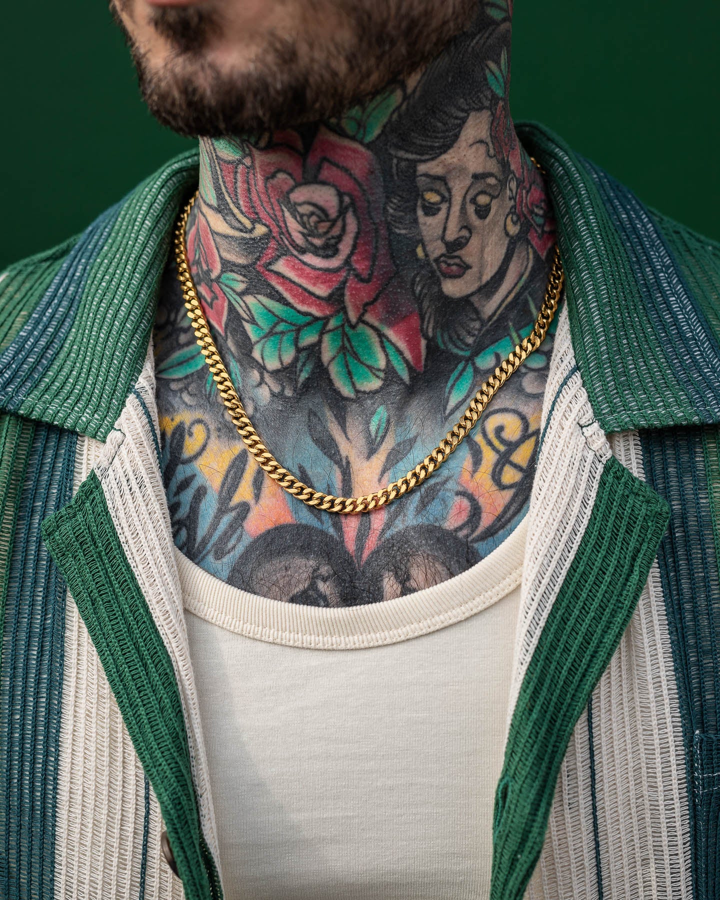A close-up of a tattooed person’s neck and upper chest, in a cream shirt and green-and-white striped jacket, showcases the eye-catching 6mm Cuban Chain (Gold).