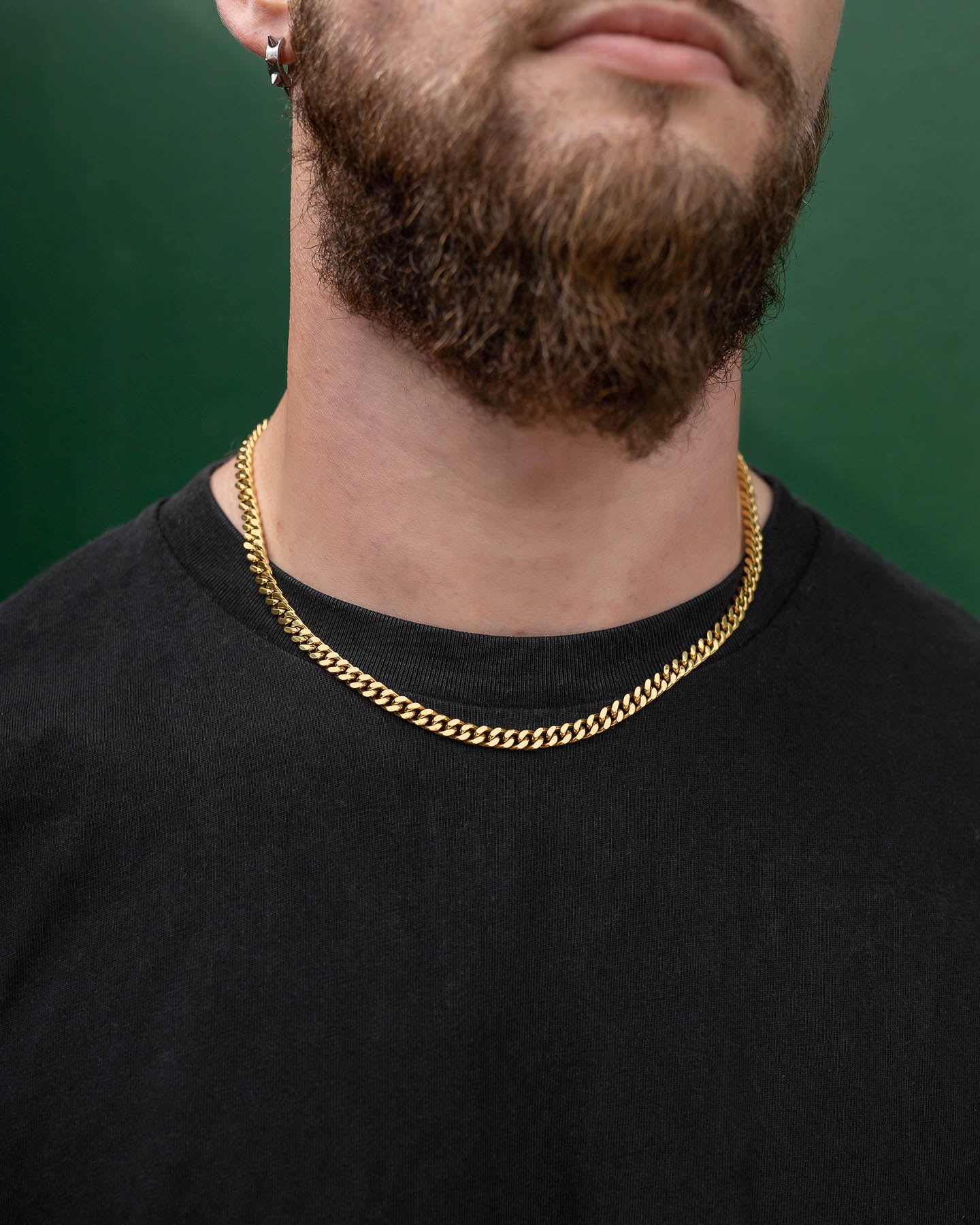 A close-up of a man wearing the 6mm Cuban Chain (Gold), a black shirt, and a small hoop earring. His lower face and beard are visible against a green background.