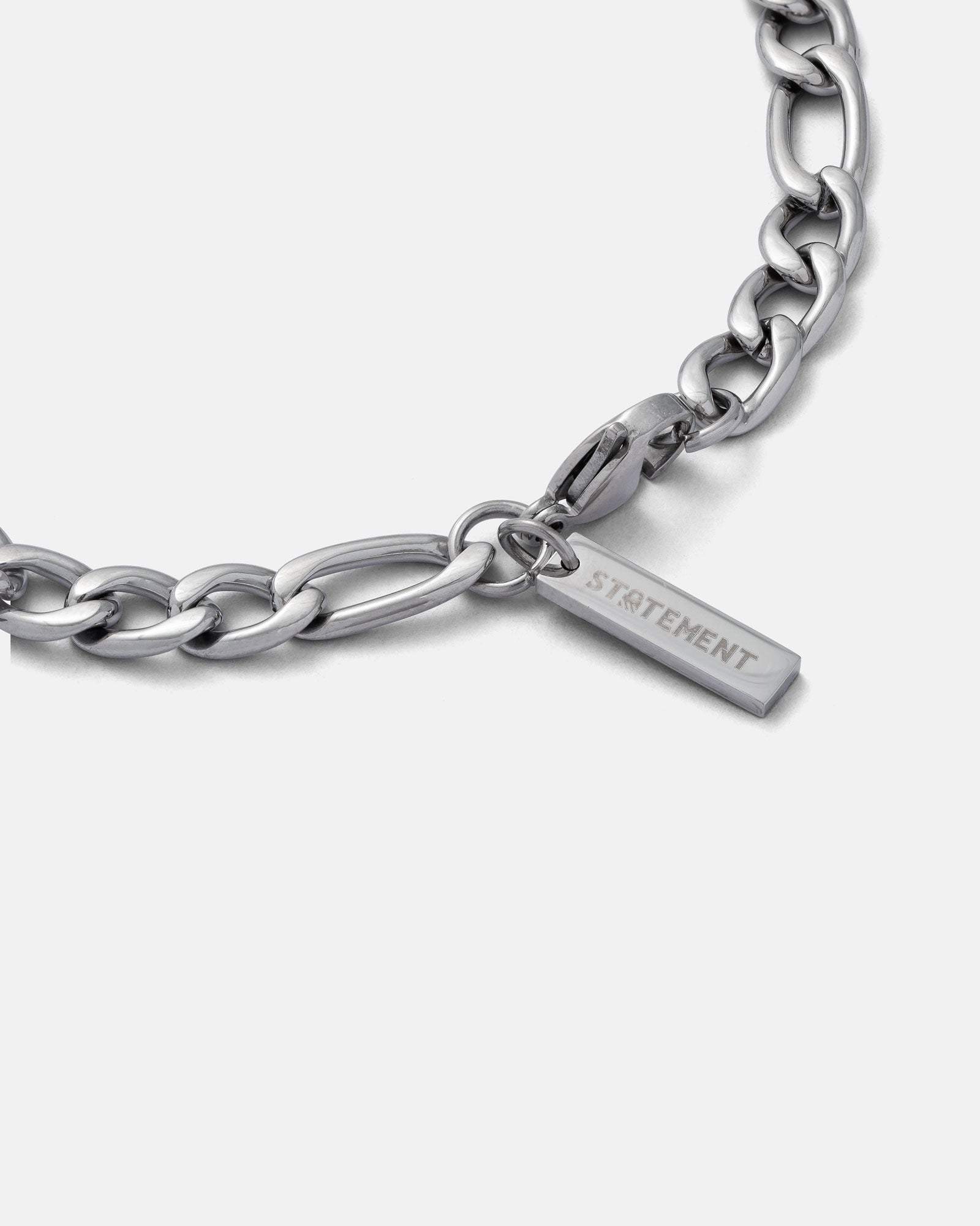 close up image of 6mm figaro chain bracelet clasp and branded tag with "STATEMENT" engraved, in silver by statement collective on grey background.
