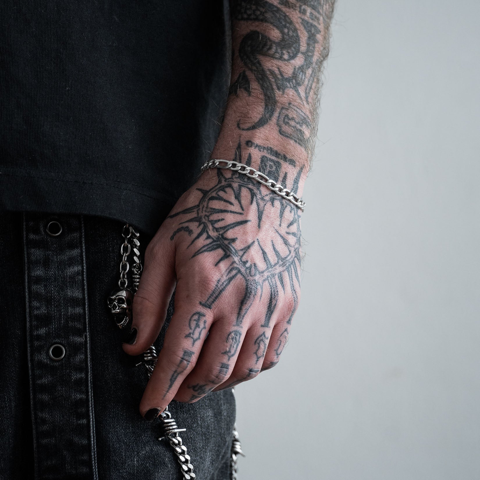 Person with tattoos on arm and hand wearing silver6mm figaro chain bracelet against a neutral background