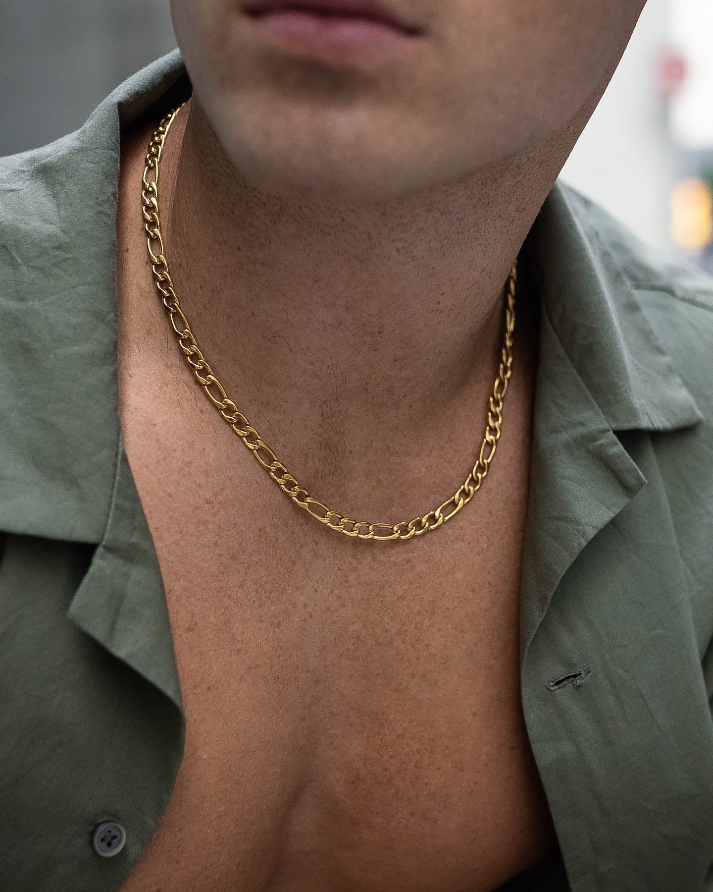 A close-up of a person wearing an unbuttoned green shirt and the 6mm Figaro Chain (Gold) necklace. The focus is on the necklace and their neck and collarbone, with their face partially out of frame.