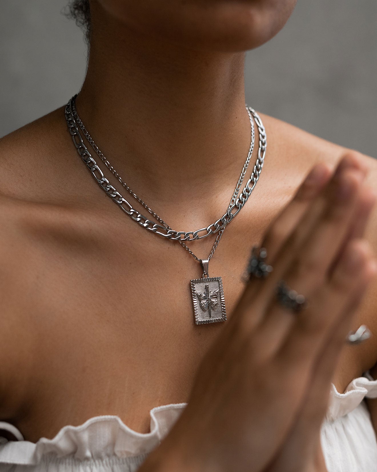 A person in a white off-shoulder top wears layered silver necklaces, including a rectangular star pendant and a 6mm Figaro Chain, plus silver rings, and holds their hands together in a prayer-like pose.