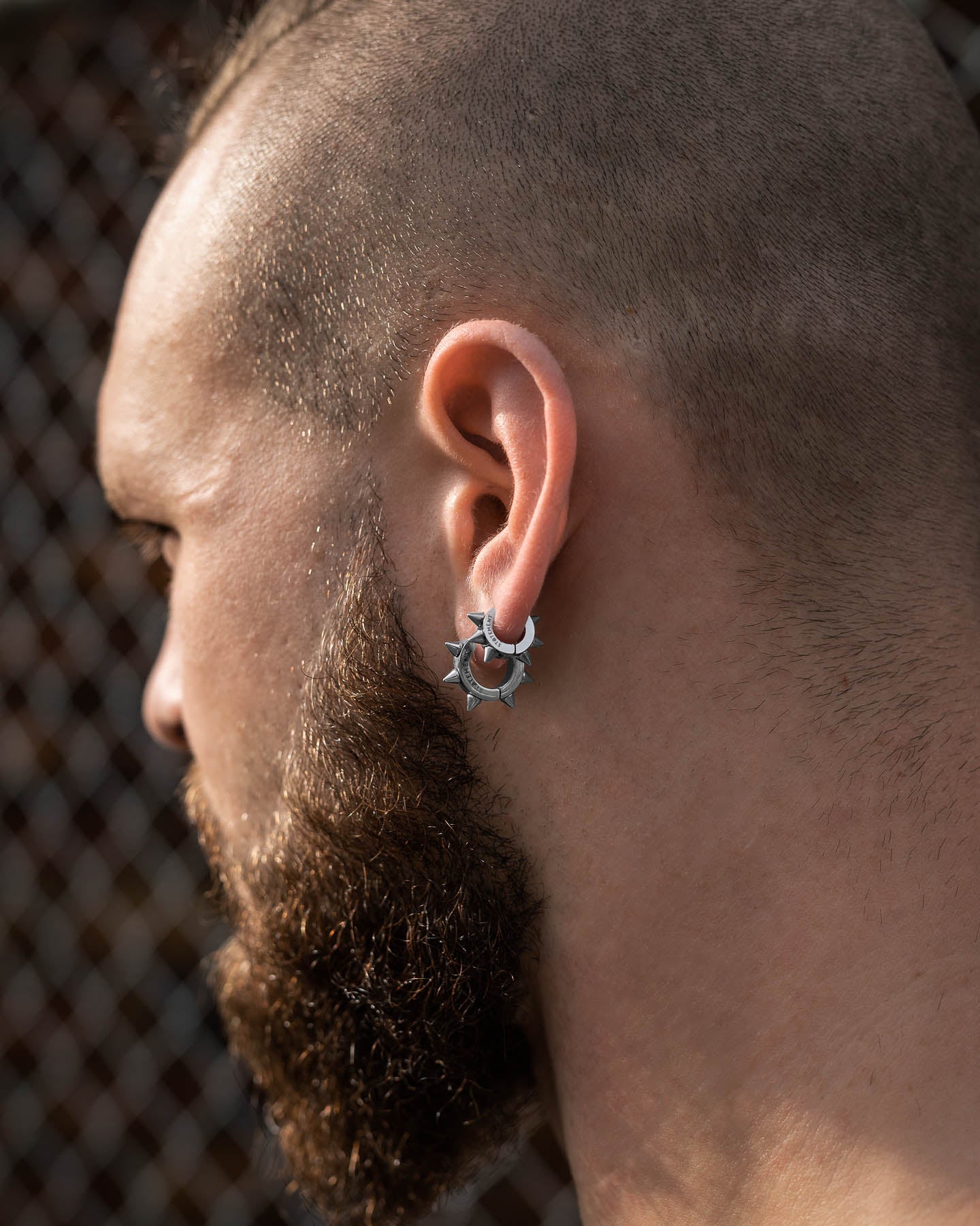 A close-up of a bearded, shaved-head person standing outdoors by a chain-link fence, wearing 6mm Spiked Hoop Earrings in their left ear.
