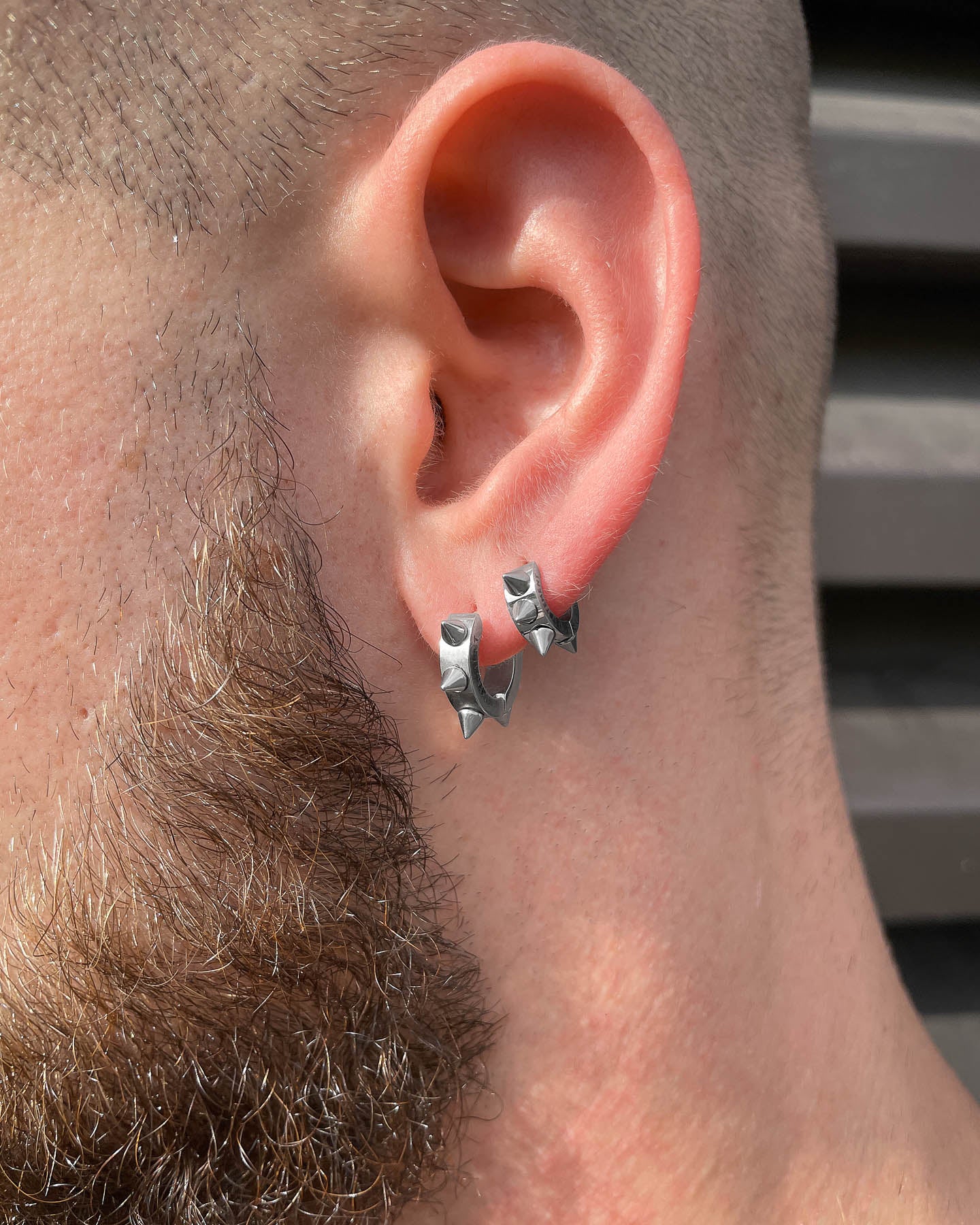 Close-up of a person’s ear wearing 6mm Spiked Hoop Earrings. The individual has a trimmed beard and short hair. The background features blurred horizontal dark lines.