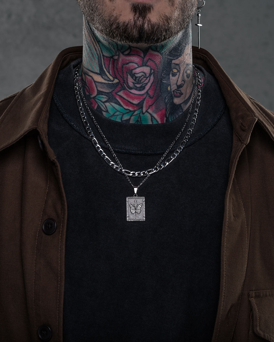 A person with colorful neck tattoos wears layered silver chains, including the 6mm Figaro Chain with a square pendant featuring a leaf design, along with a black shirt and brown jacket. Their face is partially visible.