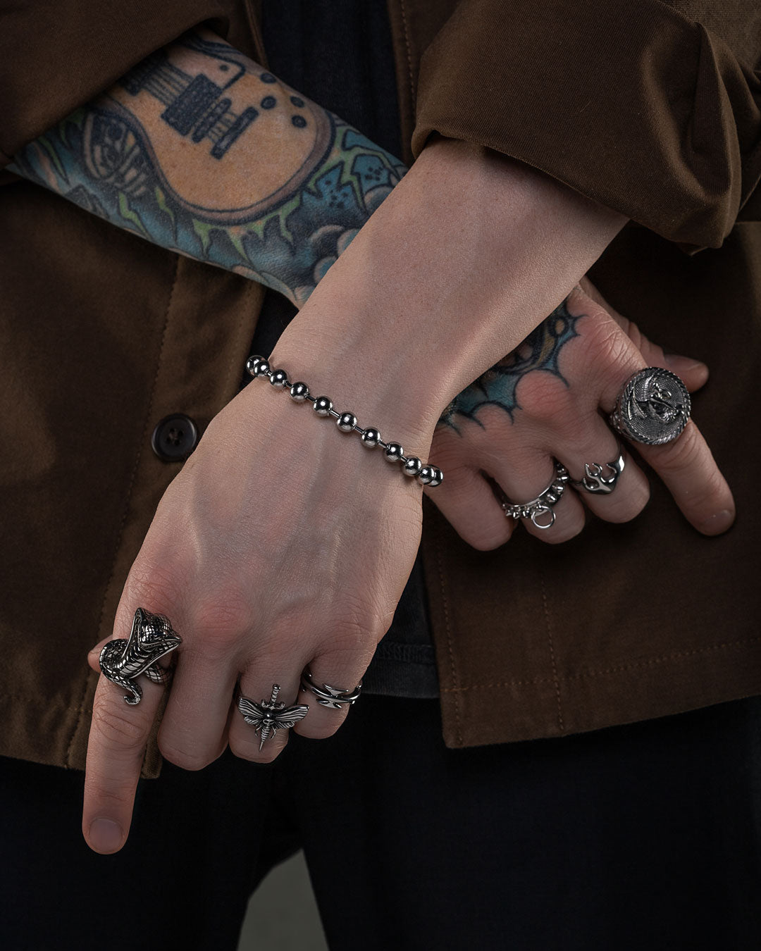 A person with tattooed hands crosses their fingers in front of a brown jacket, showcasing the 7mm Ball Bracelet alongside several silver rings for a bold, 90s-inspired vintage look.