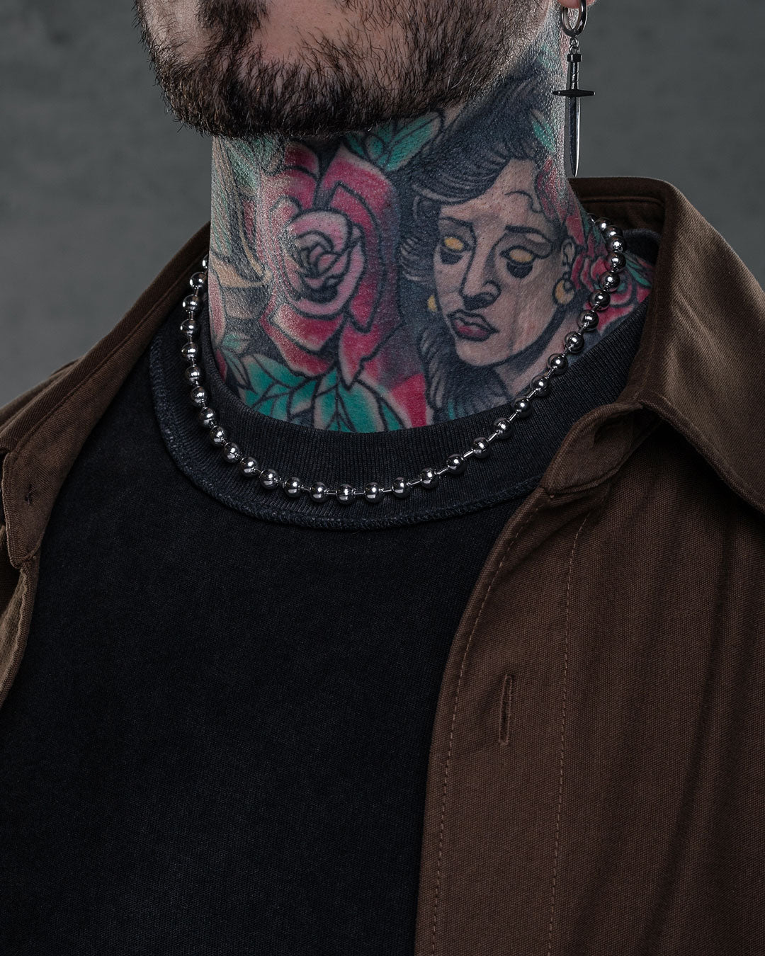 Wearing a dark shirt, brown jacket, cross earring, and the 7mm Ball Chain necklace, a person with colorful neck tattoos—featuring a rose and woman's face—is shown with only the lower half of their face visible.