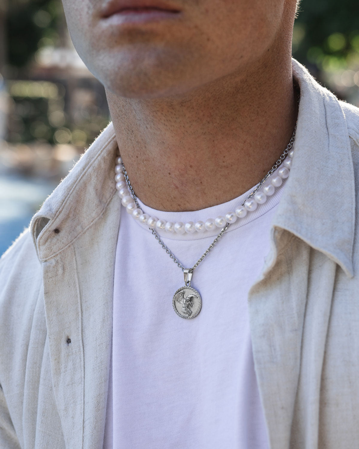 A person wearing a white T-shirt and beige shirt, layered with the 8mm Freshwater Pearl Necklace and a silver chain with an engraved round pendant. The image highlights the neck and chest area.