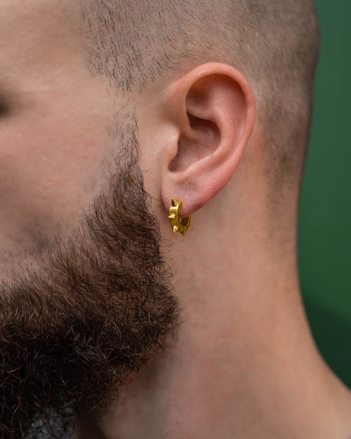 Close-up of a person's ear wearing 8mm Spiked Hoop Earrings (Gold), highlighting their edgy punk style. The individual has a full brown beard, short hair, and is set against a vibrant green background.