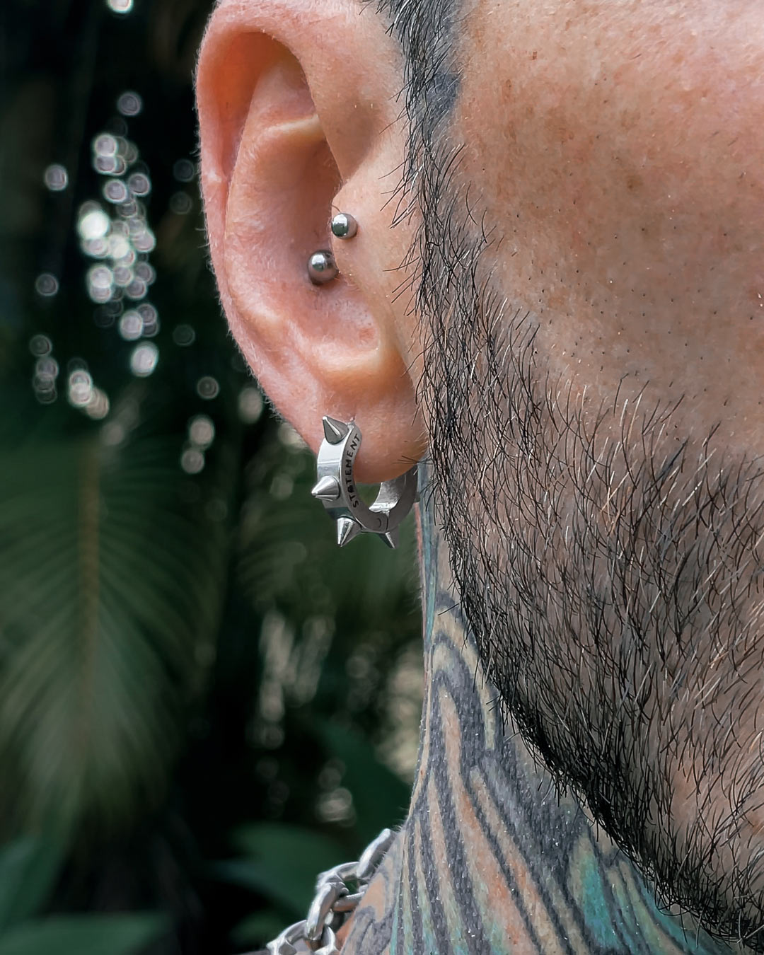 Close-up of a person’s ear wearing 8mm Spiked Hoop Earrings in the lobe and a silver piercing on the upper ear; a tattooed neck and stubbled cheek are also visible, with blurred greenery in the background.