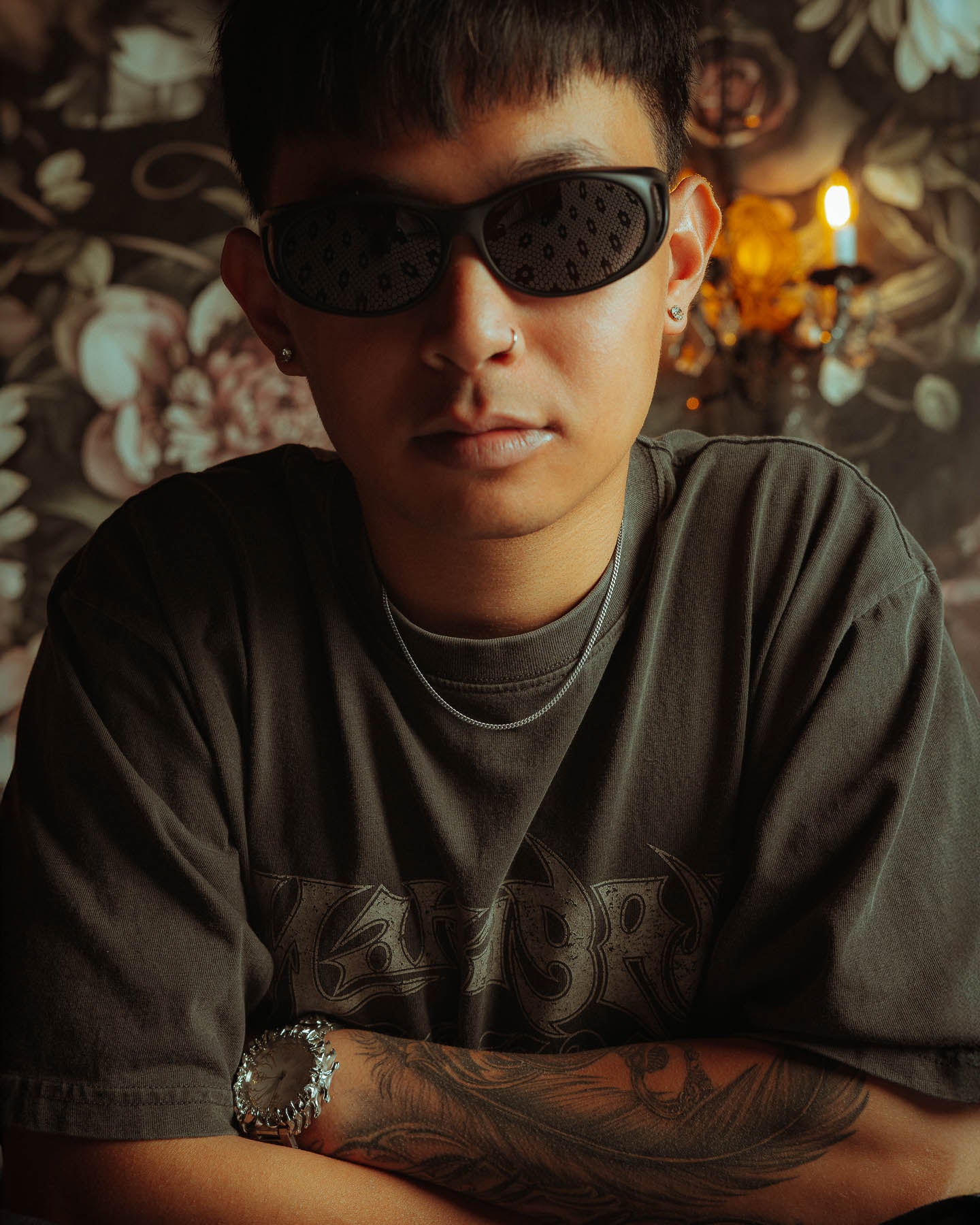 A person with short dark hair, black sunglasses, a graphic tee, wristwatch, and tattooed arm poses with crossed arms wearing a 925 Sterling Silver 2.3mm Curb Chain in front of a floral wall and lit sconce.