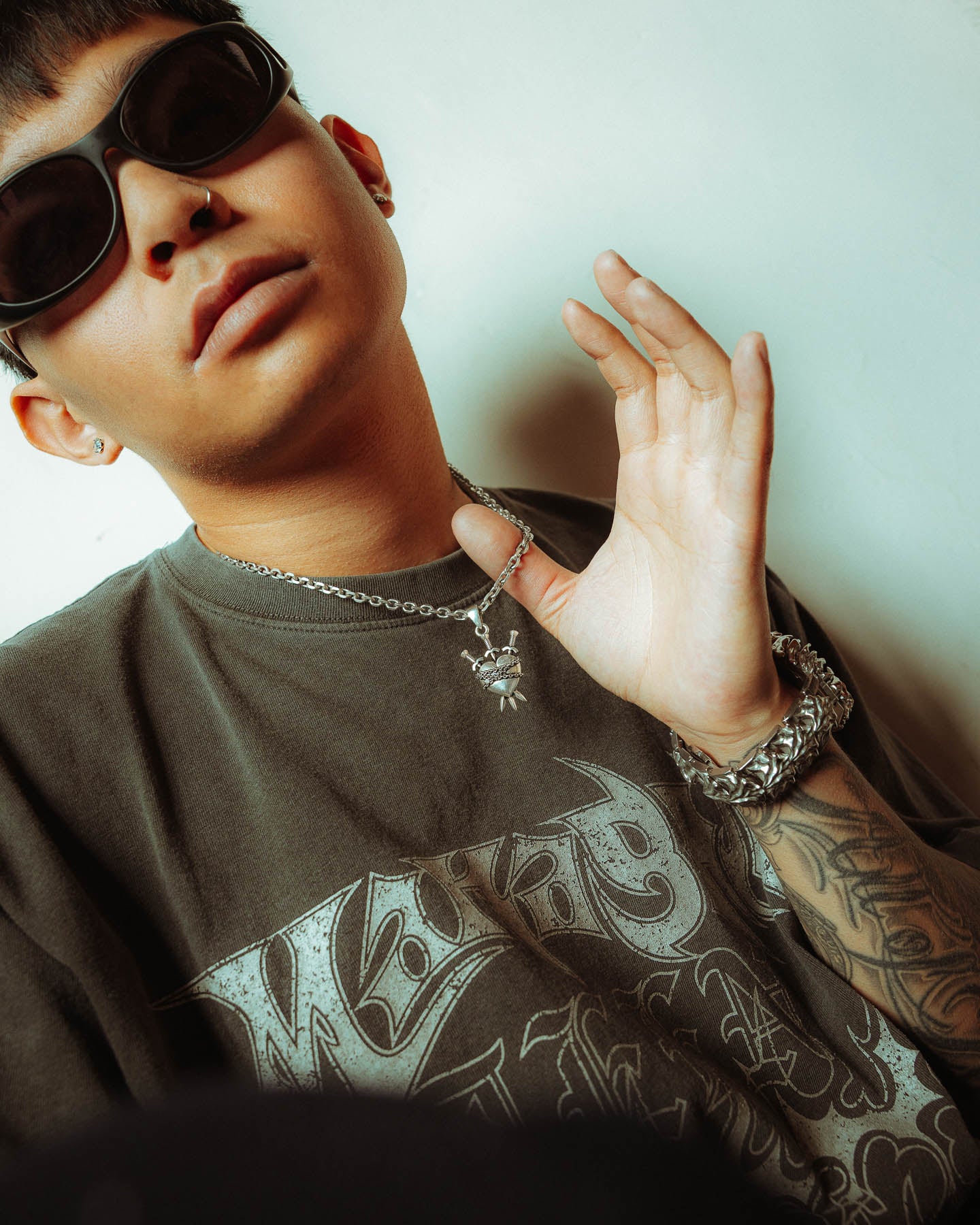 Wearing dark sunglasses, a graphic tee, jewelry—including a chain bracelet and the 925 Sterling Silver "3 of Swords" Pendant—a person poses with one hand raised near their tattooed arm, calmly looking at the camera.
